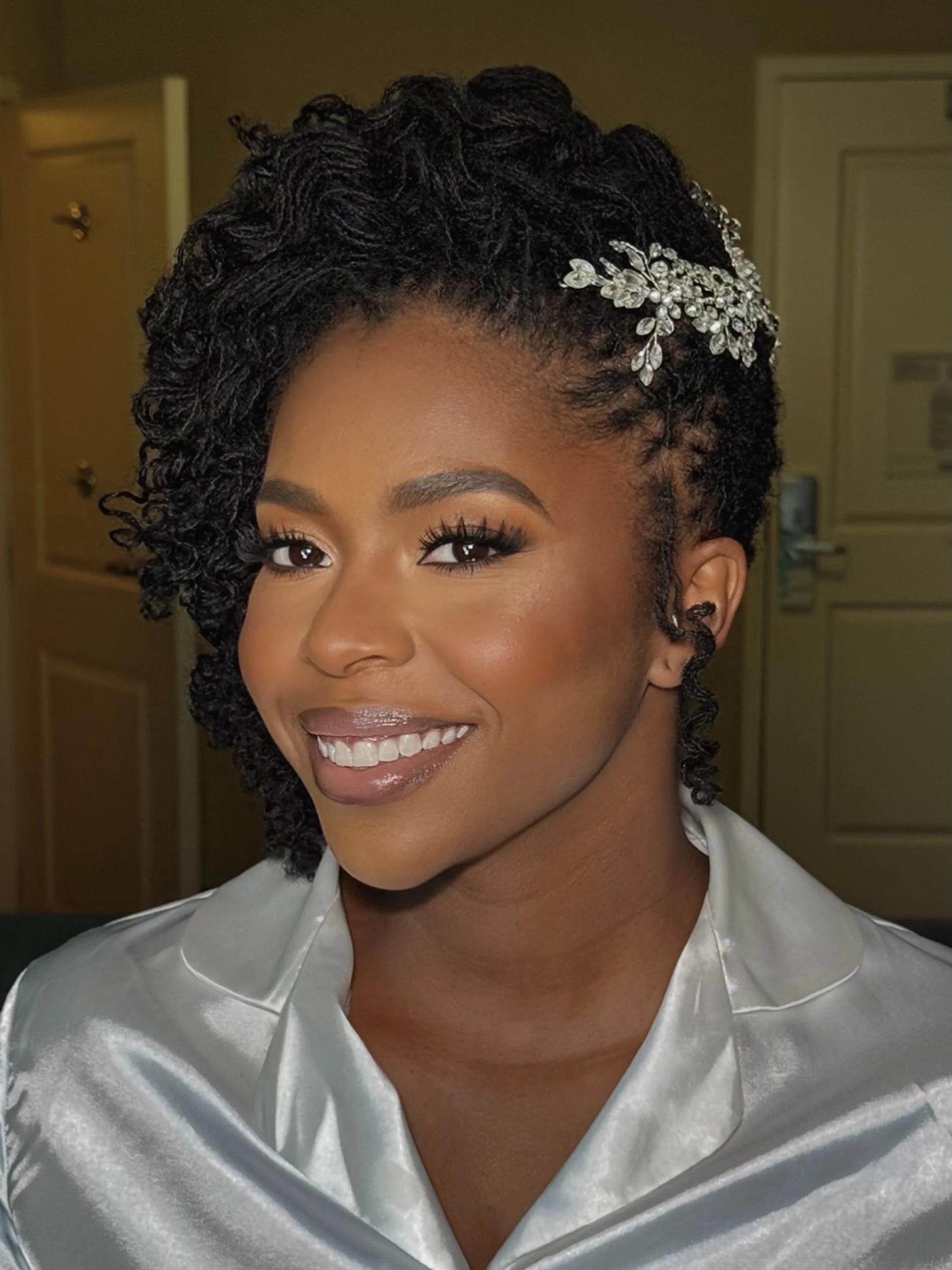 A woman with styled curly hair wearing a decorative hairpiece, smiling at the camera, dressed in a shiny satin robe.