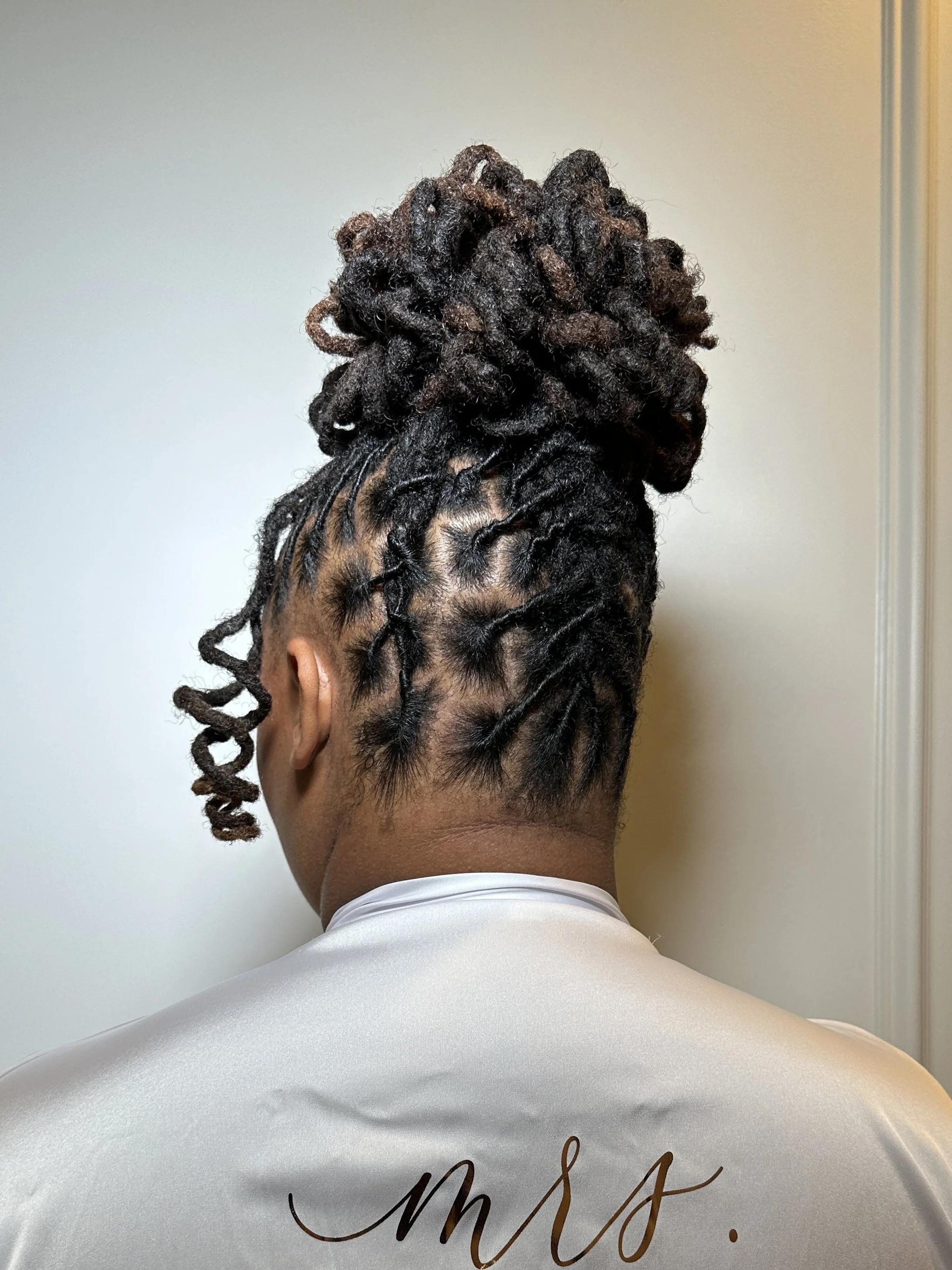 Back view of a person with short, styled dreadlocks in dark brown and blonde shades tied up in a high bun, wearing a white shirt with cursive writing on the back.