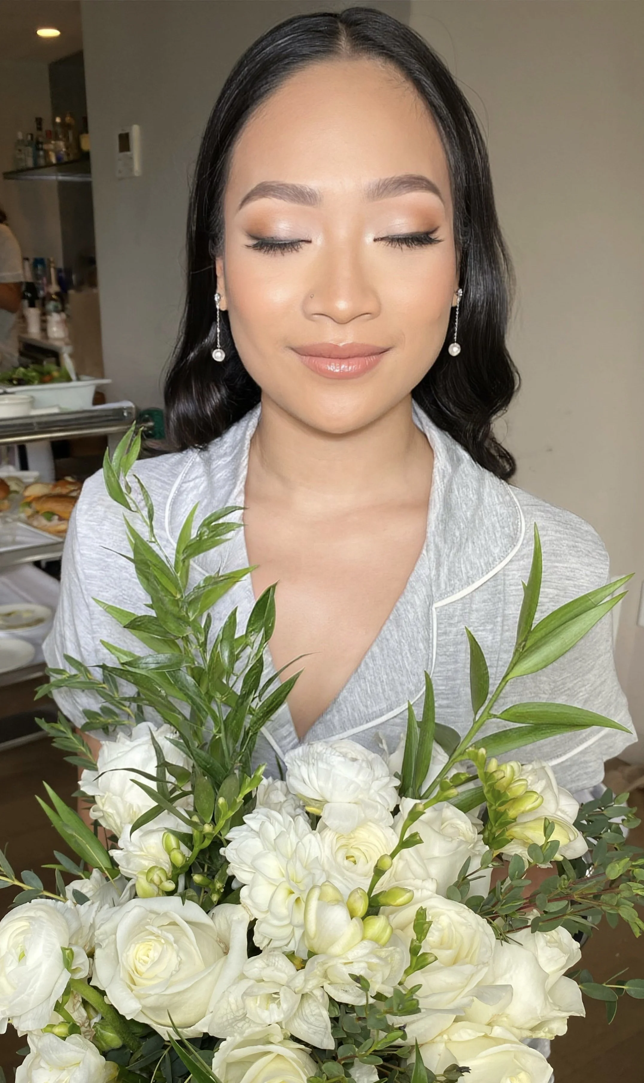 A woman with light makeup and pearl earrings is holding a large bouquet of white flowers, including roses and lilies, with a neutral background.