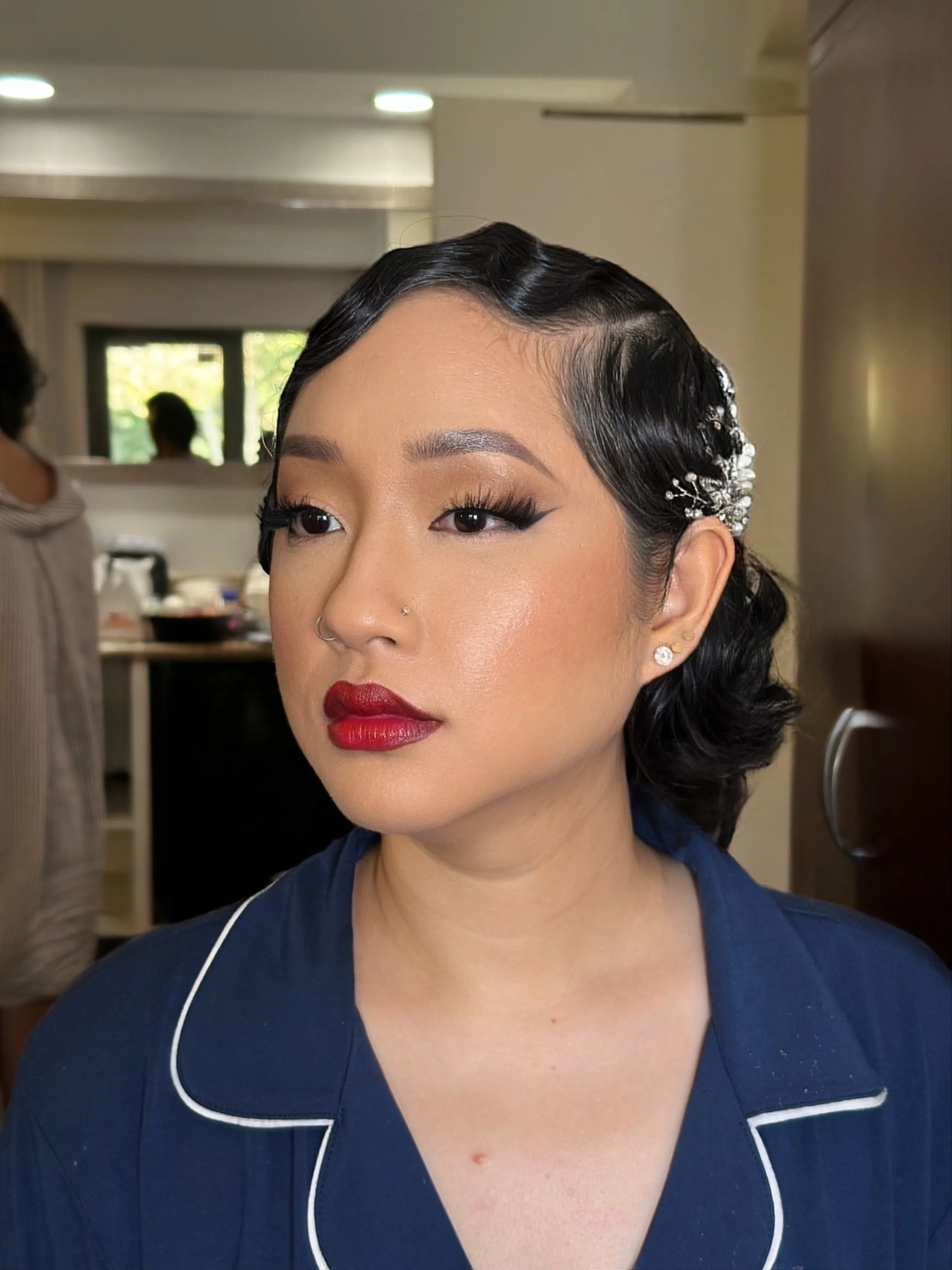 A woman with vintage styled black hair accessorized with a decorative hairpiece, wearing red lipstick, eyeliner, and diamond earrings, dressed in a navy-blue uniform with white piping, indoors with a blurred background.