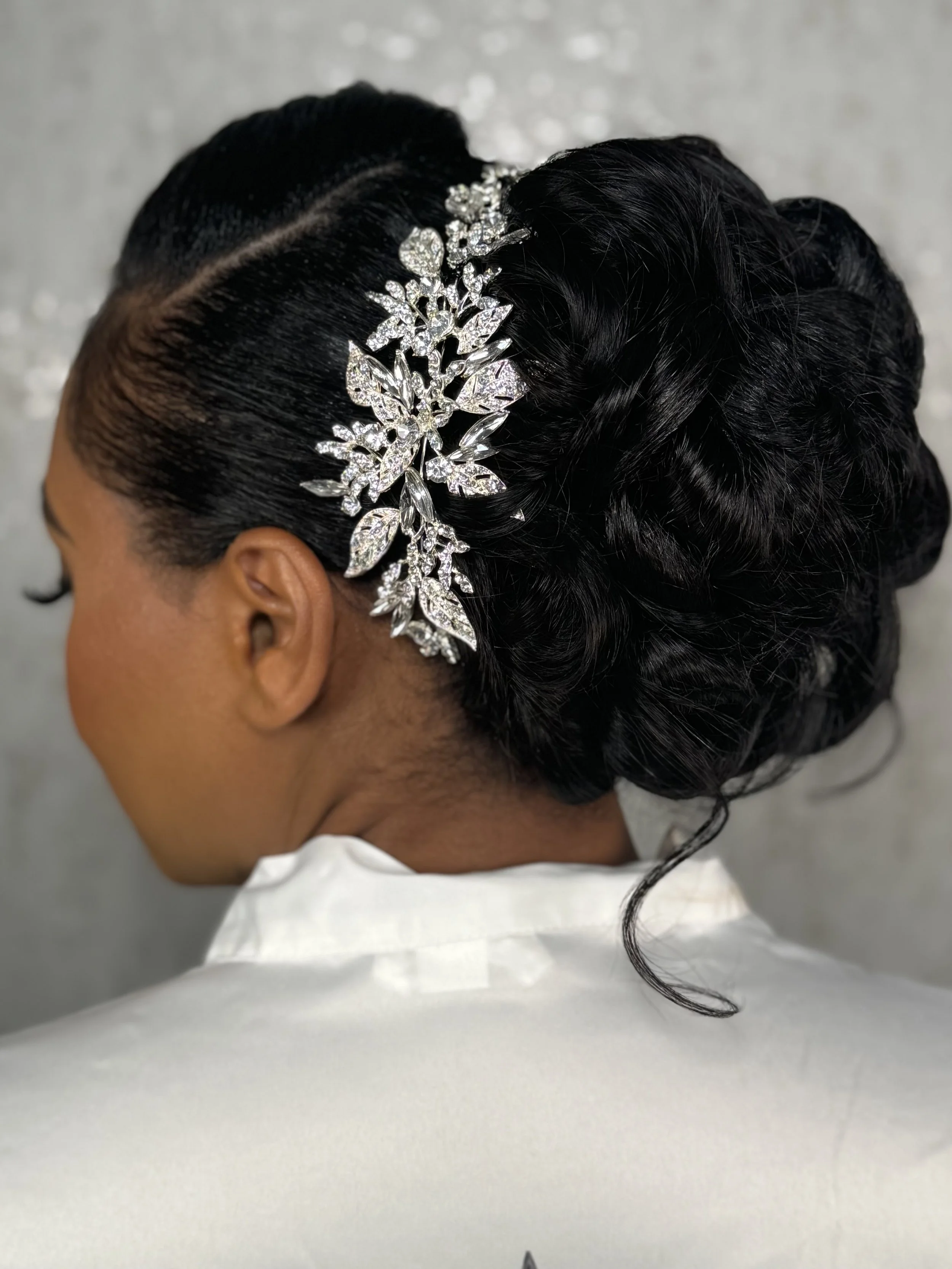A close-up of a woman with dark, styled hair adorned with a sparkling leaf and floral hair accessory, wearing a white outfit, viewed from the side.