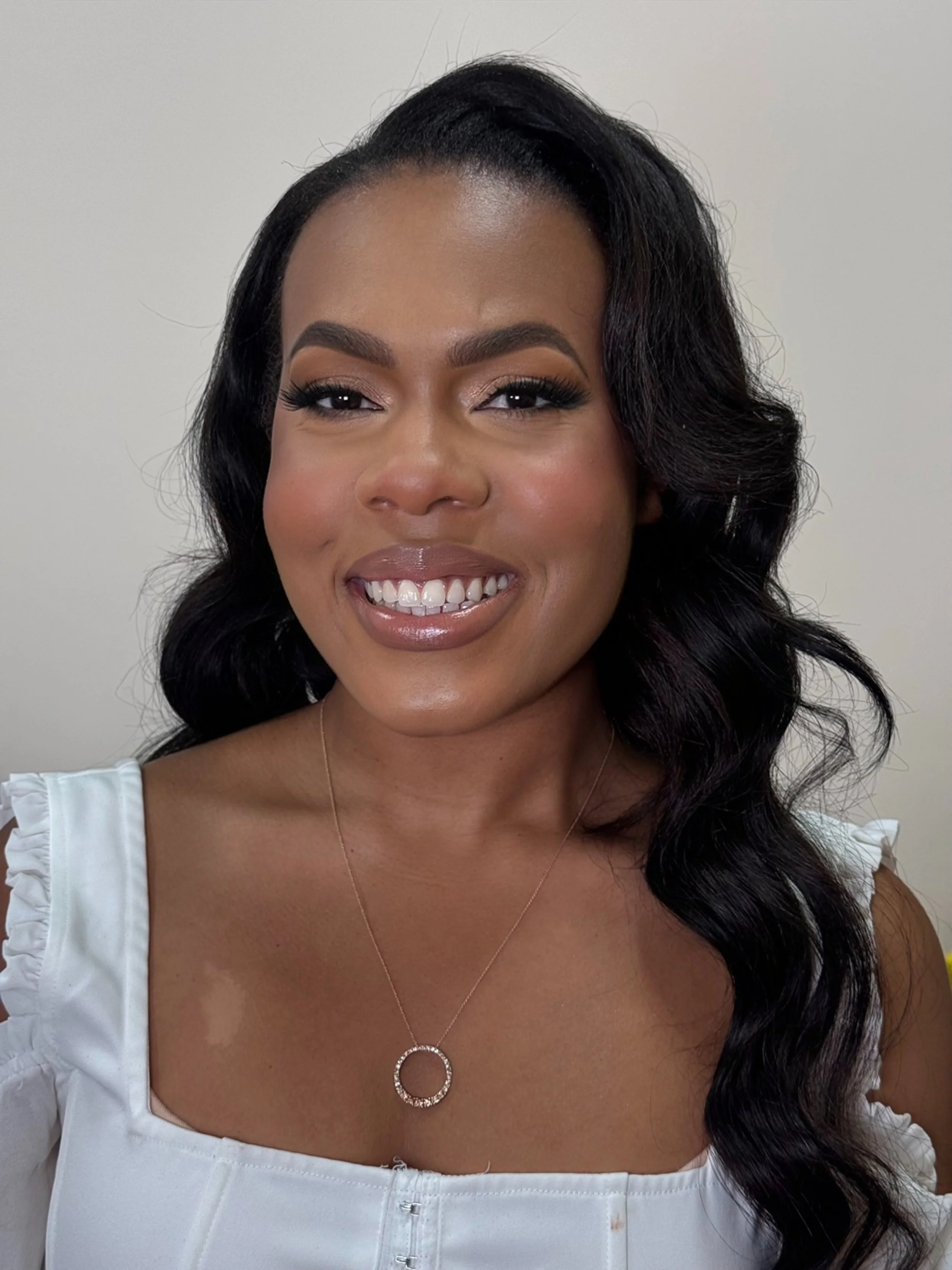 Close-up of a smiling woman with dark wavy hair, wearing a white top with ruffled sleeves and a silver necklace with a circular pendant.