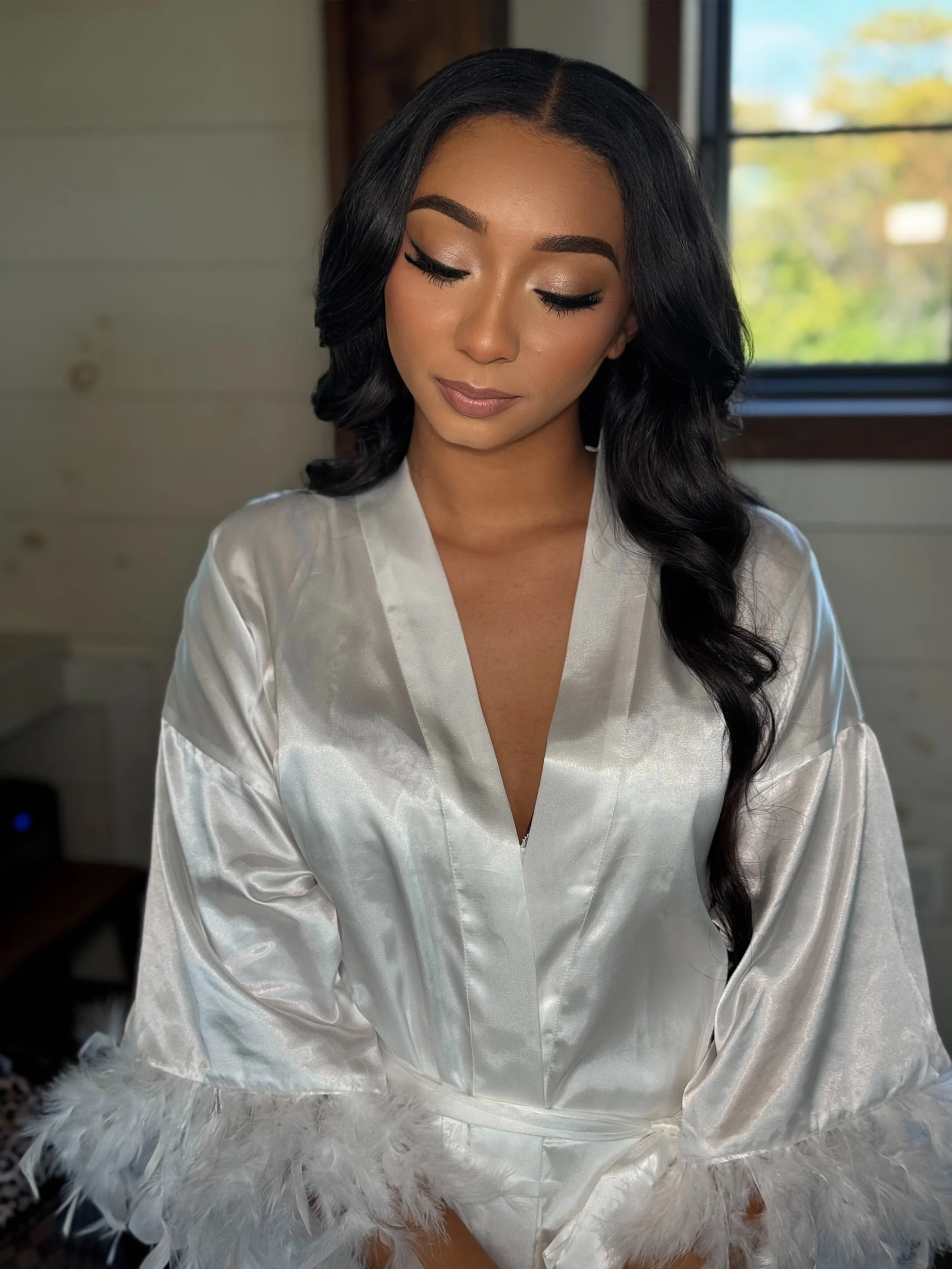 A woman with long black hair and makeup wearing a satin white robe sitting on a fluffy white surface in front of a window with trees outside.