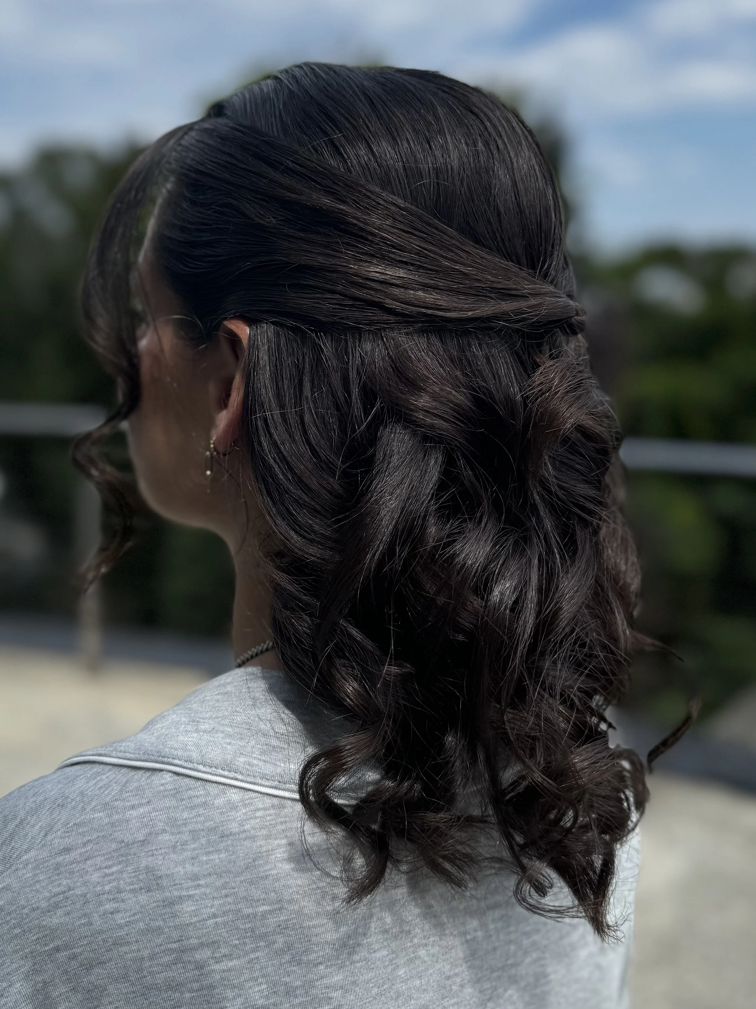 Close-up of a woman with dark brown, curly hair, wearing a gray shirt and small earrings, outdoors on a cloudy day.