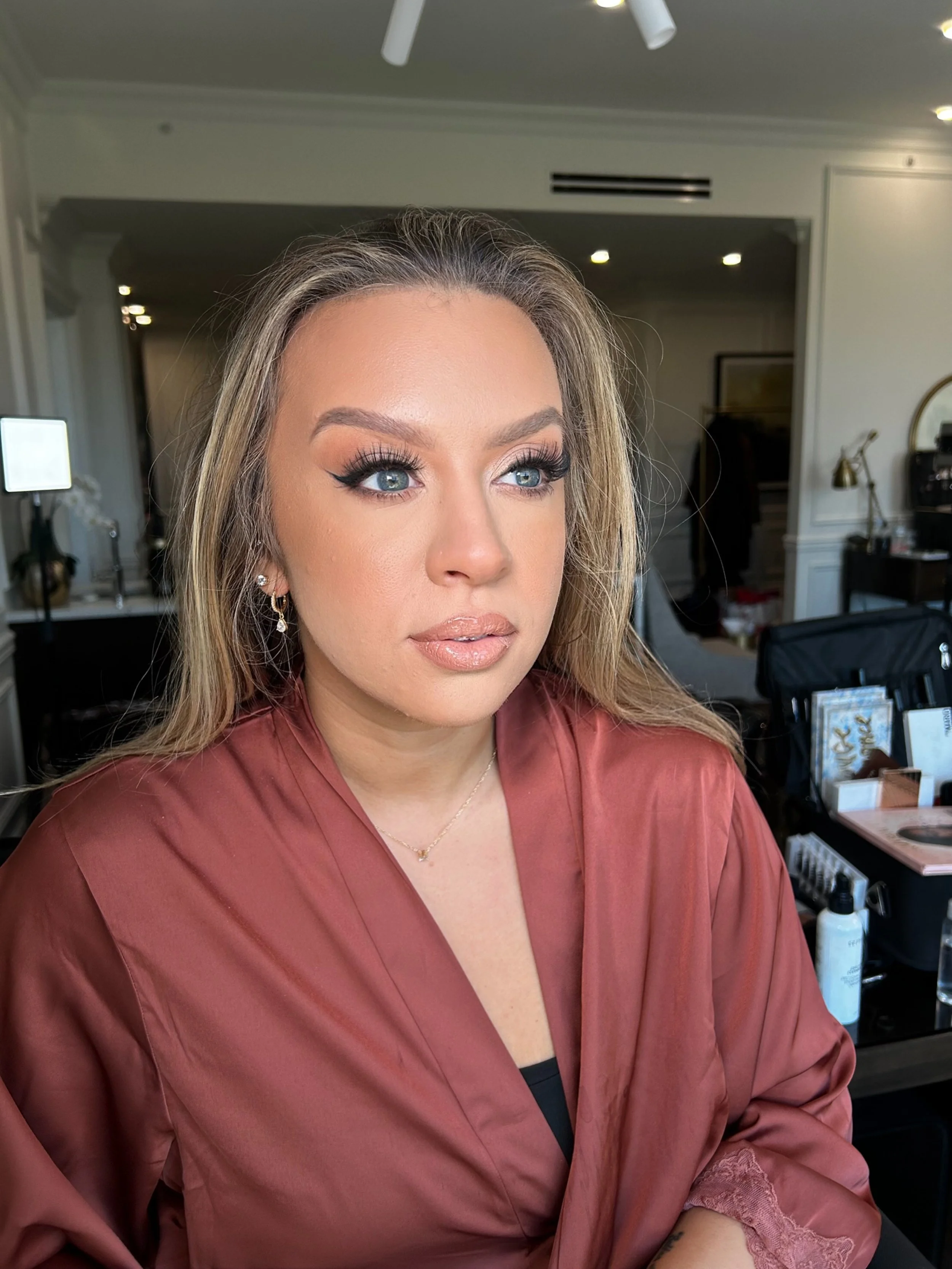 Close-up of a woman with blonde hair, blue eyes, and makeup, sitting in a room with makeup and beauty products in the background.