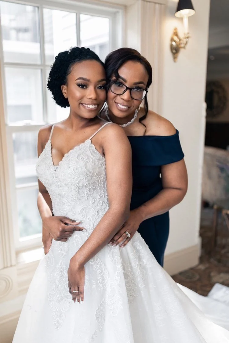 Two women, one in a wedding dress and the other in a formal dress, smiling and posing together indoors near a window.