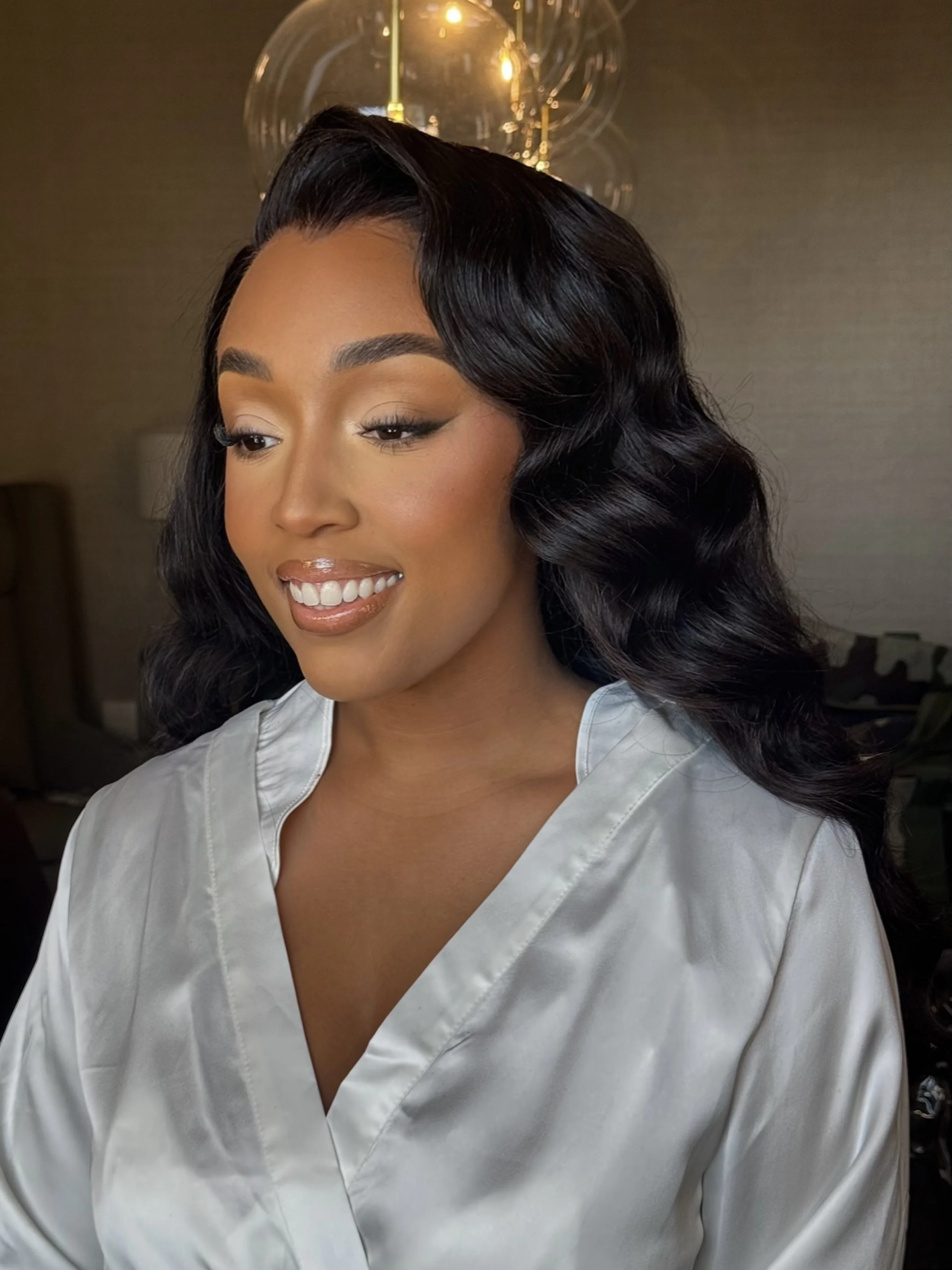 A woman with medium brown skin, long black wavy hair, smiling and wearing a satin white robe, stands in a room with beige walls and a modern hanging light fixture in the background.