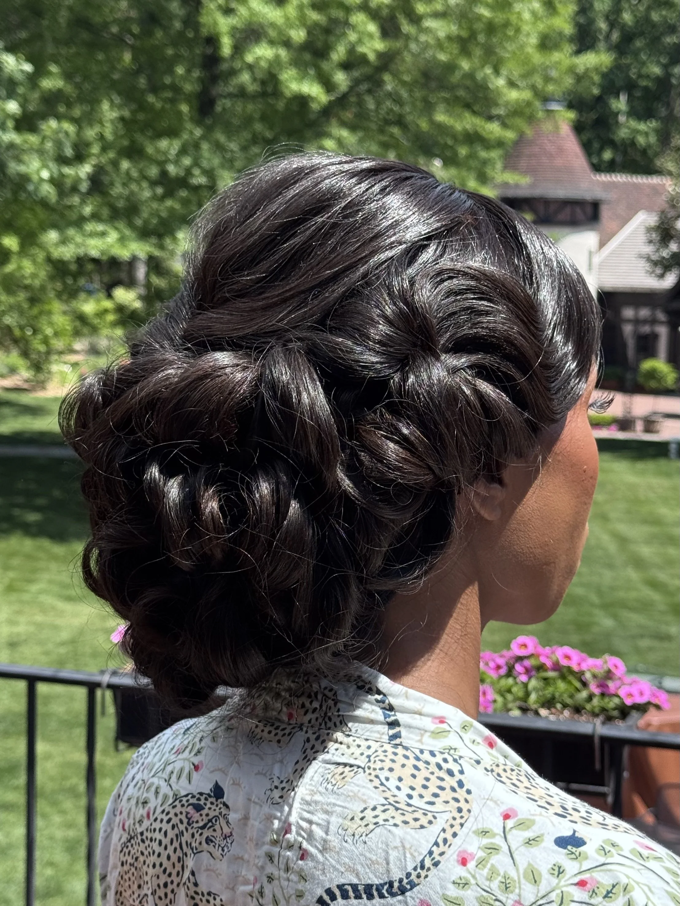 Close-up of a woman with dark, shiny, curly hair styled in an elegant updo, seen from the side against a background of green trees, a house with a thatched roof, and pink flowers.
