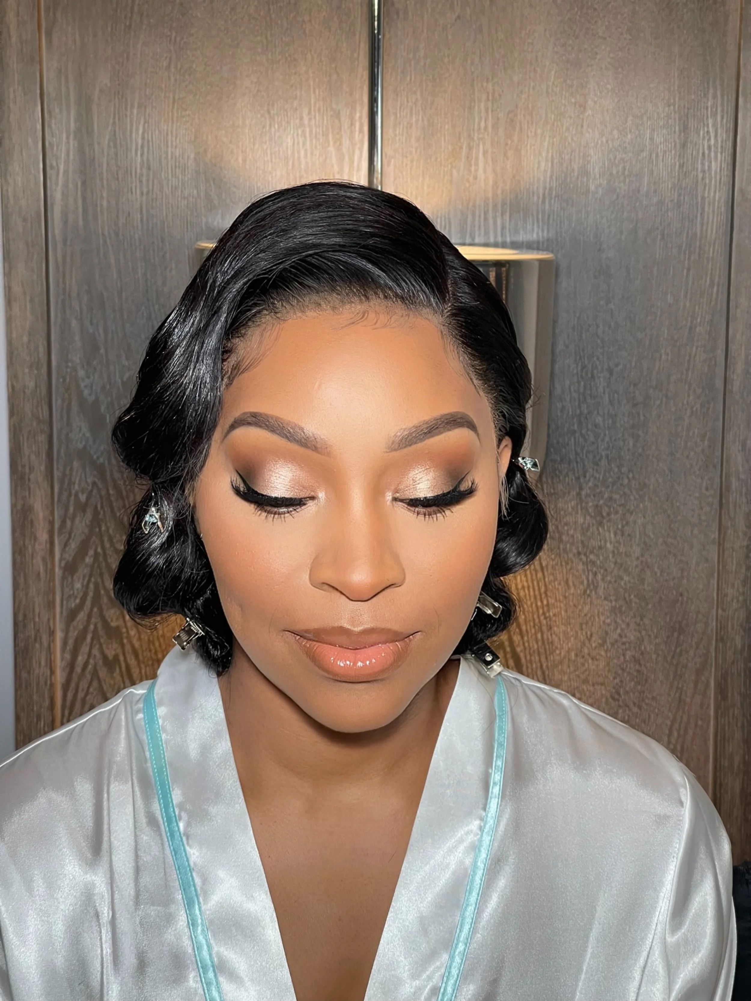 A woman with styled black hair and makeup, wearing a satin robe with light blue trim, with a neutral facial expression and closed eyes.