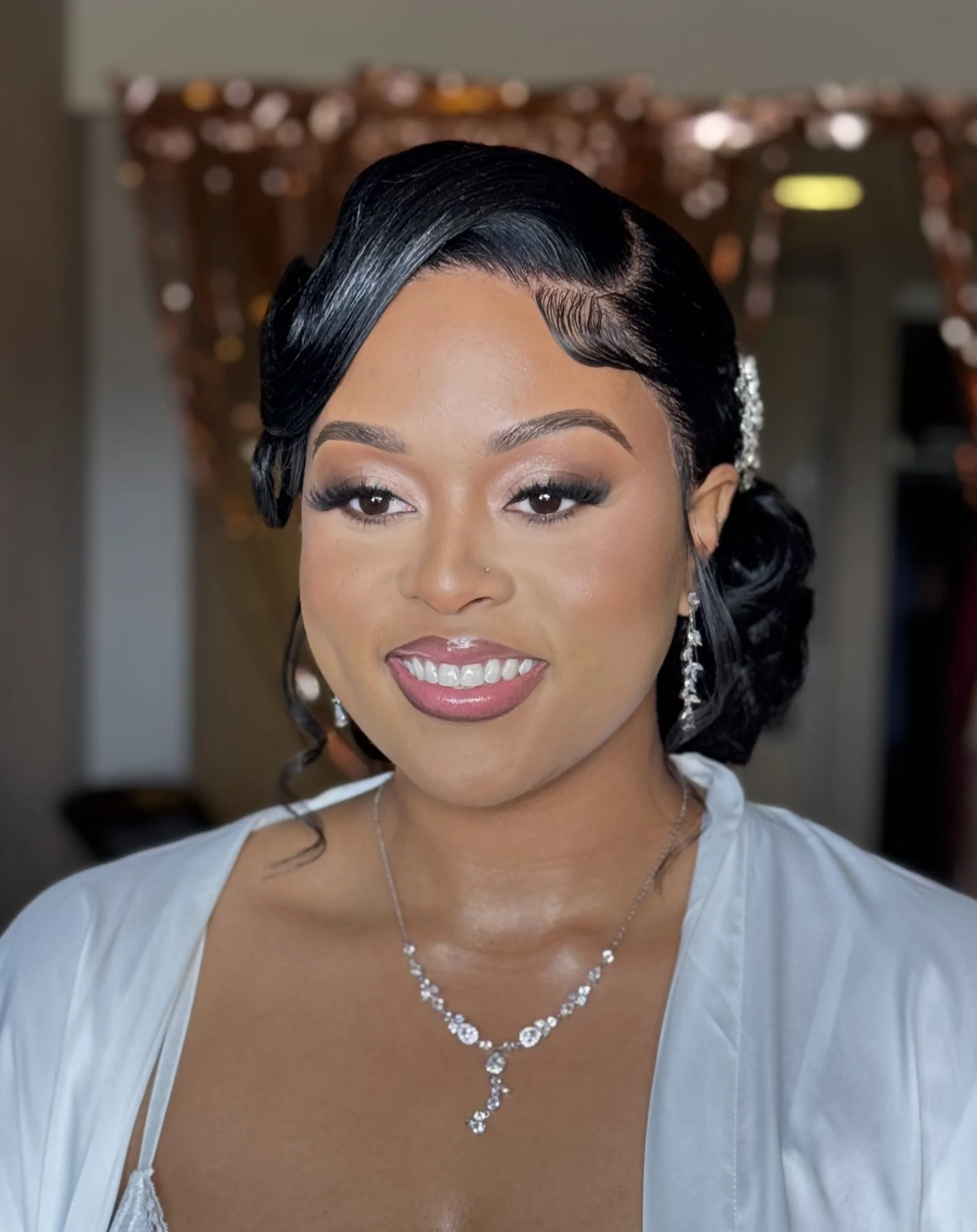 Close-up of an woman with black, styled hair, makeup, and jewelry, smiling, with a blurred background.