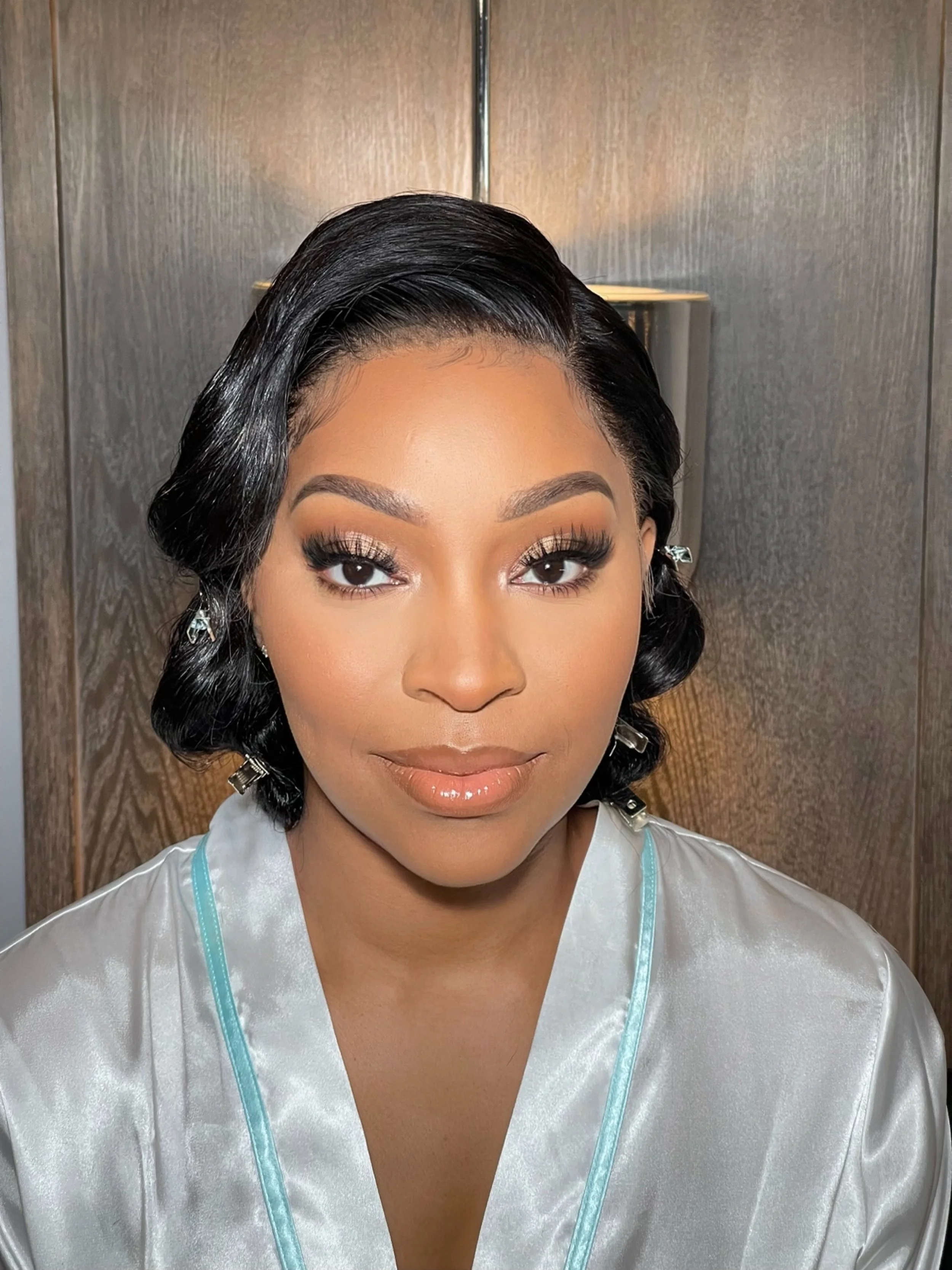 Close-up of a woman with styled black hair, makeup including eyebrows and eyelashes, wearing a satin white robe with teal trim, standing against a wooden background.