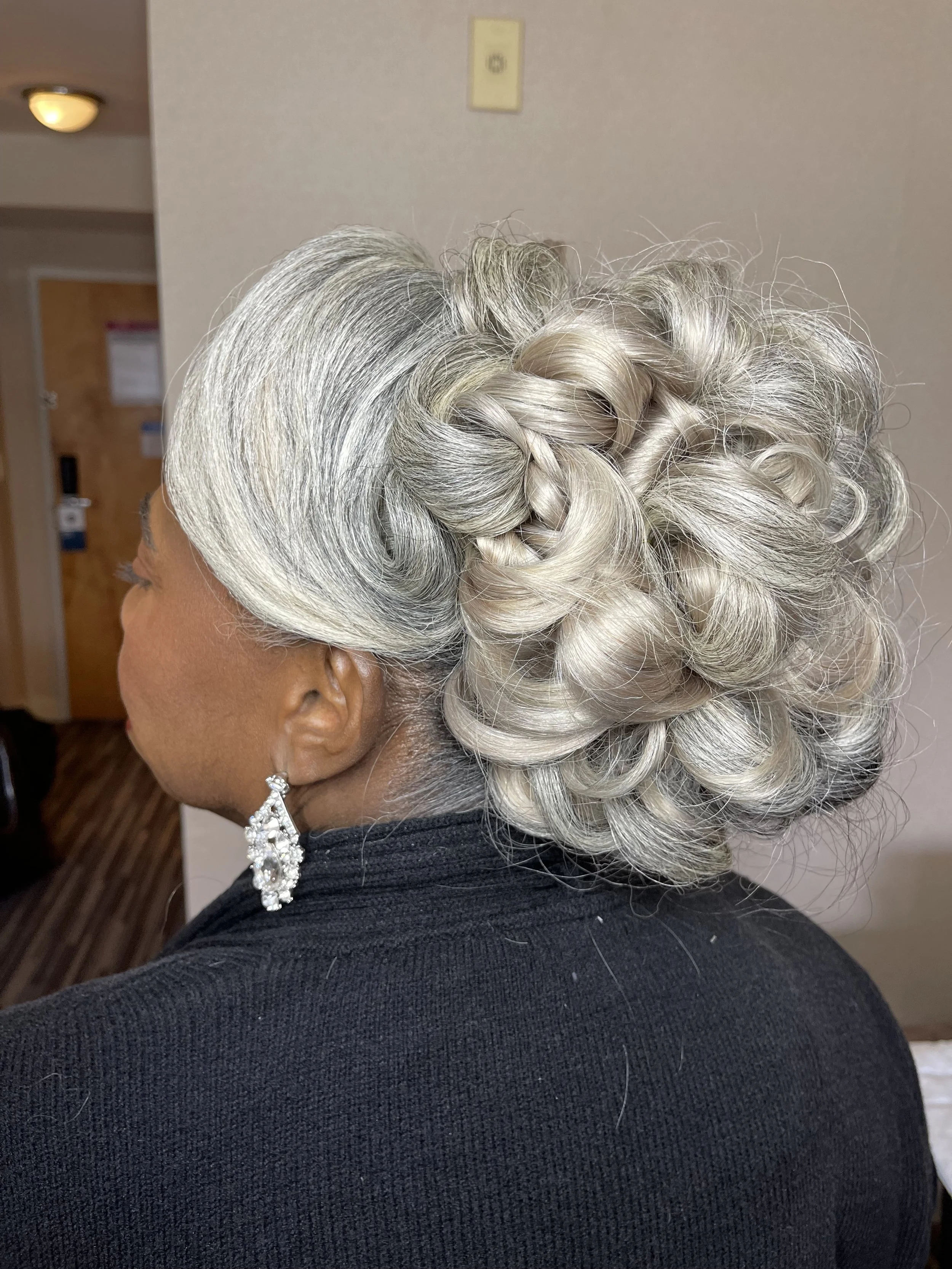 Close-up of an elderly woman's head showcasing a glamorous silver and white curly updo hairstyle with a braid, wearing a black top and large dangling earrings.
