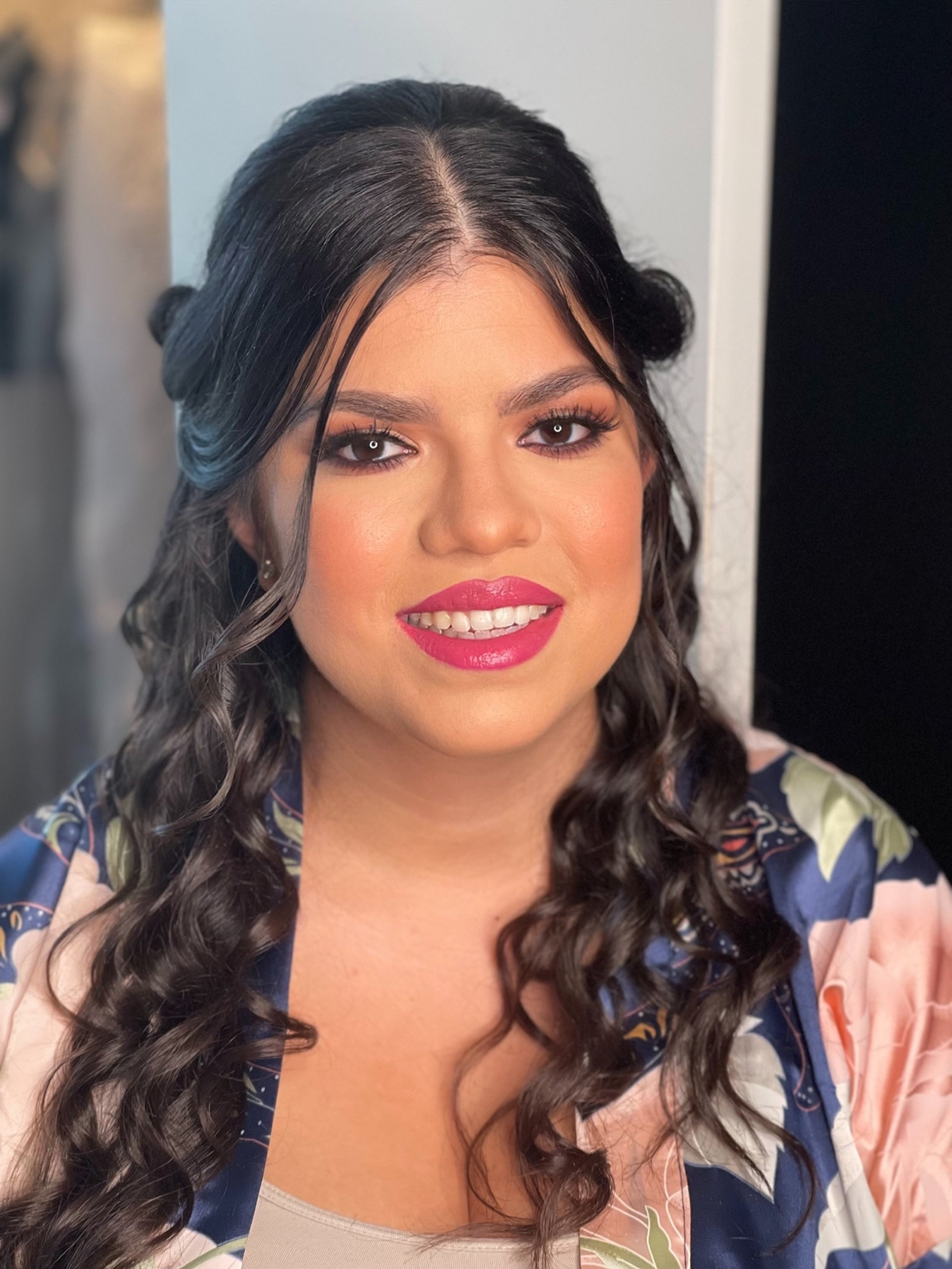 A woman with dark, curly hair styled with loose waves, wearing makeup including dark eyeliner, brown eyeshadow, and bright pink lipstick, smiling at the camera. She is dressed in a colorful, patterned blouse with floral designs.
