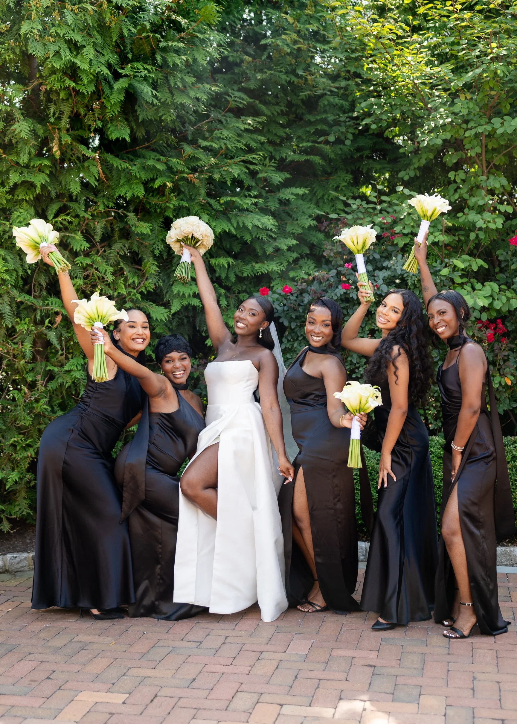 Group of six women in formal attire celebrating outdoors with bouquets, smiling, and raising their flowers in the air, standing on a brick pathway with green bushes and trees in the background.