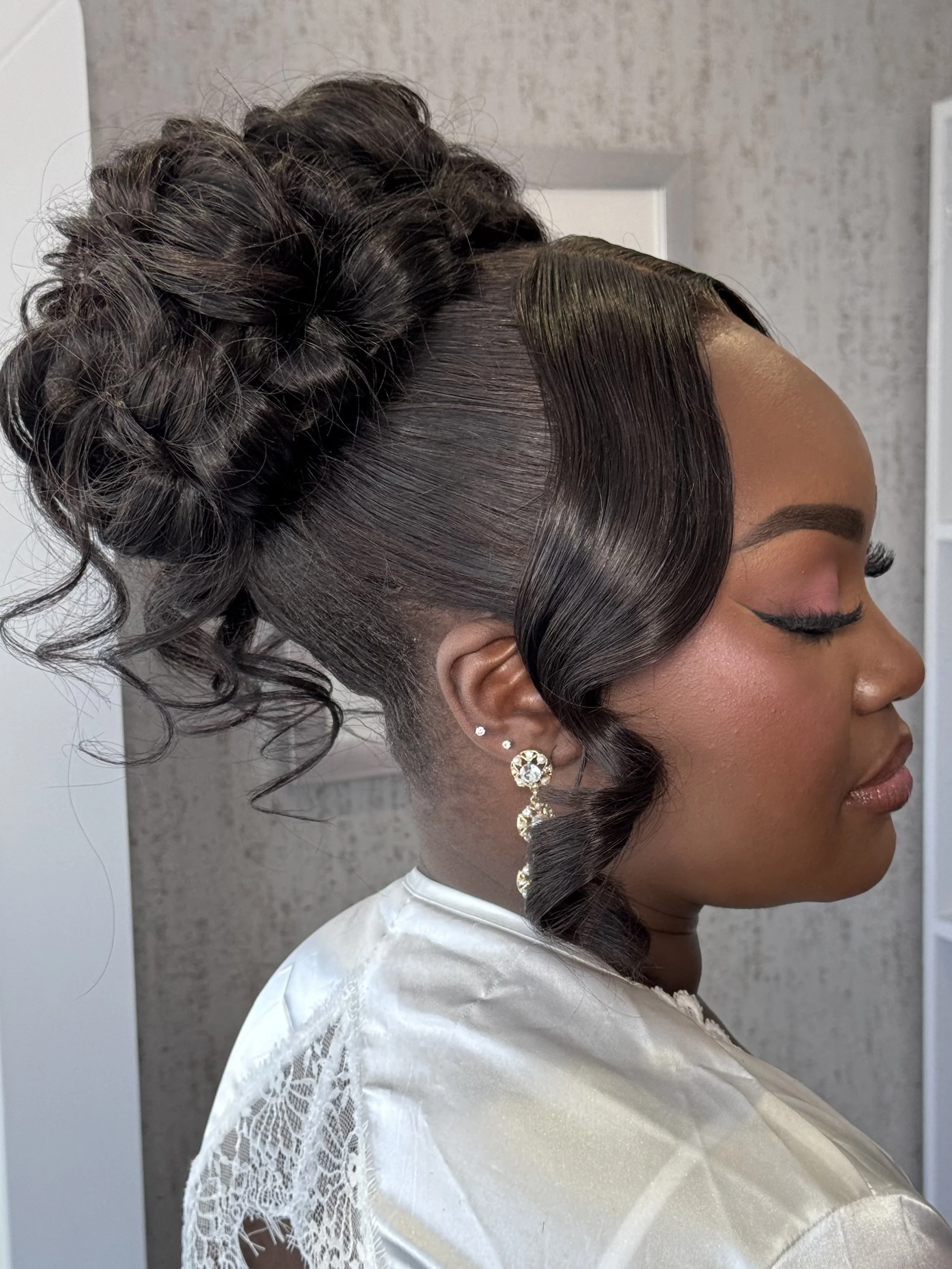 A woman with elegant makeup and jewelry, with her hair styled in an updo featuring side-swept curls and a braided crown.