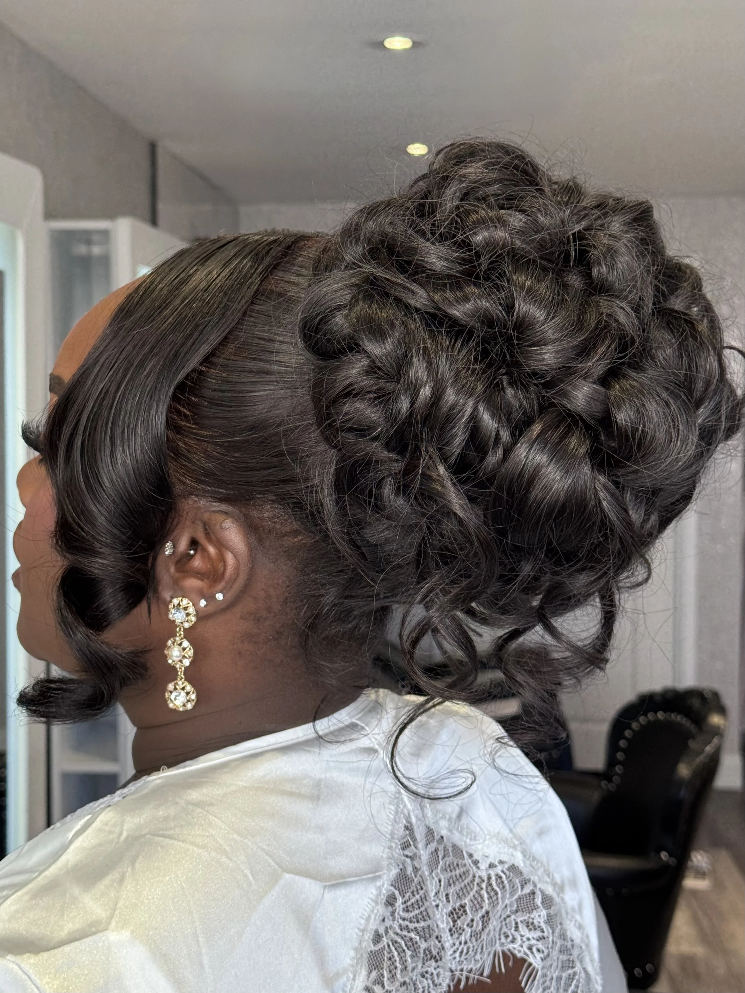 A woman with curly hair styled in an updo, wearing large pearl and rhinestone earrings, dressed in a cream-colored satin outfit with lace details.