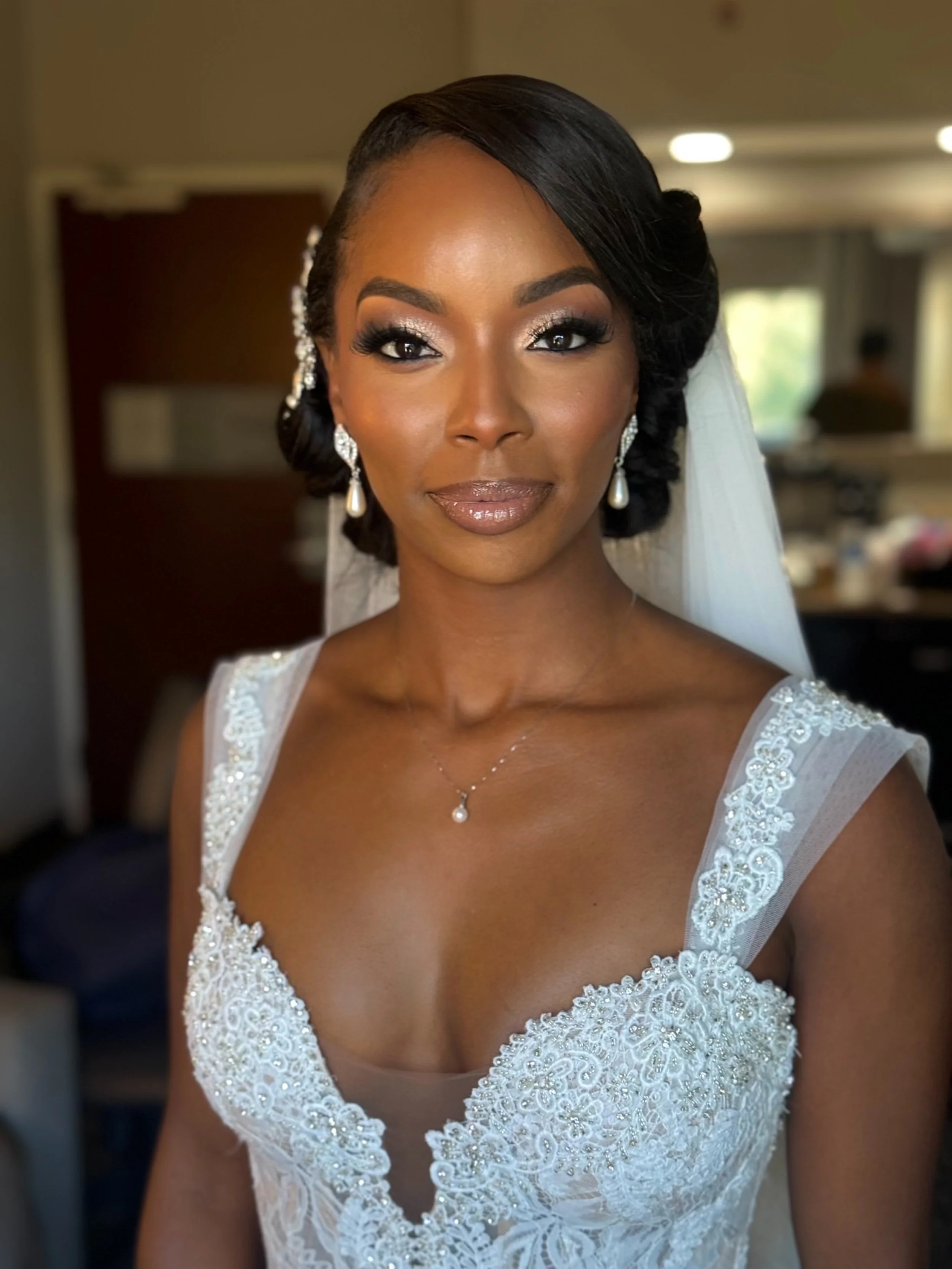 A close-up portrait of a bride with dark hair styled in a vintage wave, wearing pearl earrings, a delicate necklace with a pearl pendant, and a white lace wedding dress with detailed embroidery.