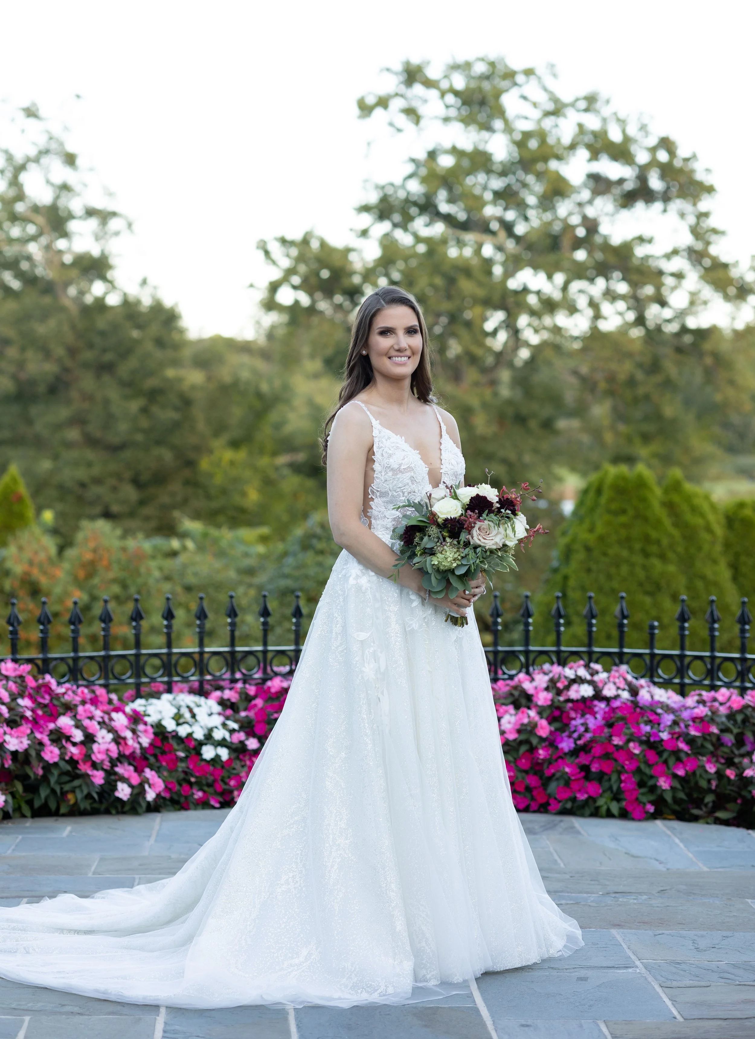 A bride in a white wedding gown holding a bouquet of flowers outdoors in front of a colorful flower bed and green trees.