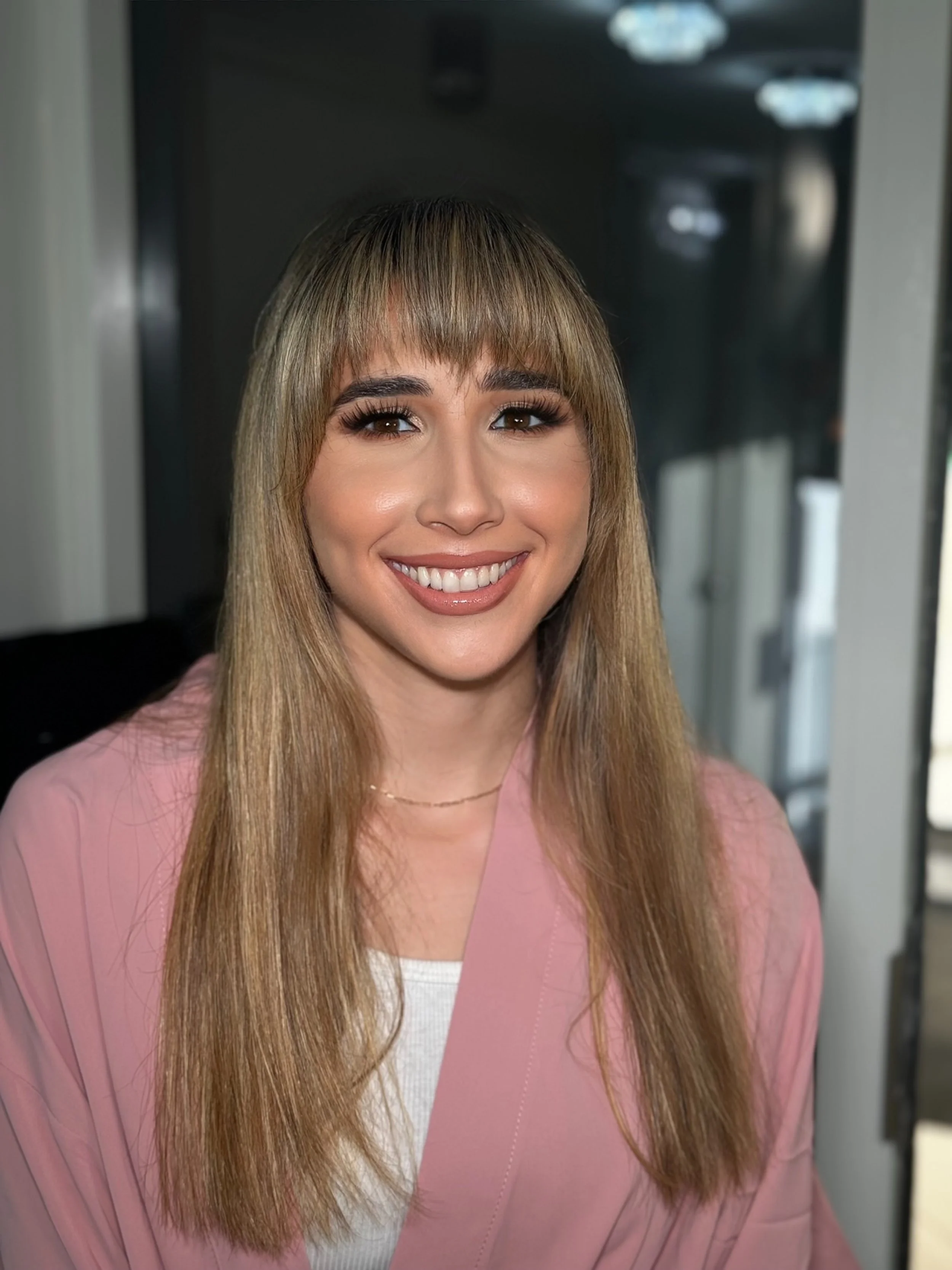 A smiling woman with long blonde hair and bangs, wearing a pink blazer over a white top, standing inside a building with modern interior and soft lighting.