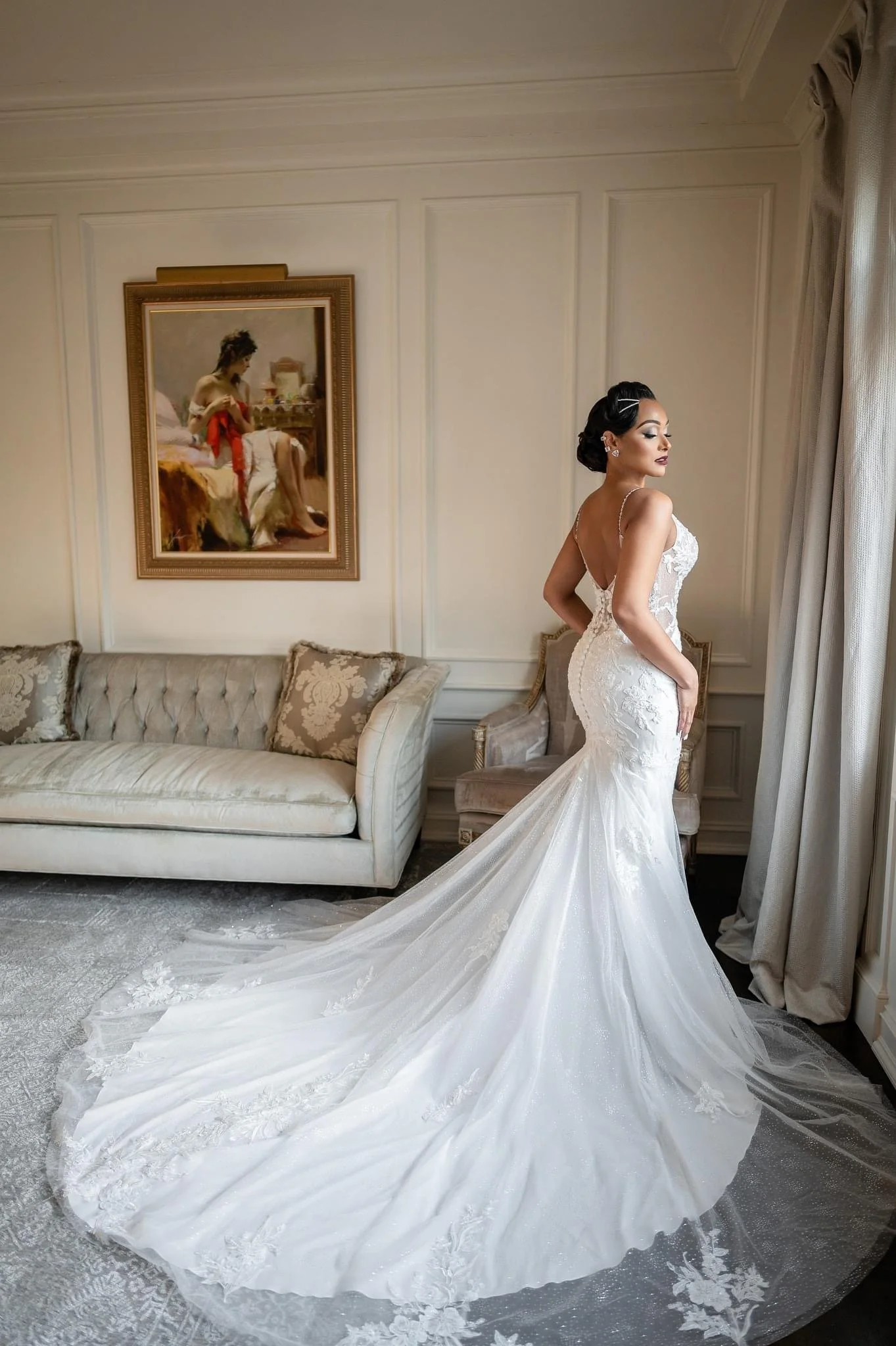 A bride in a white wedding gown with lace details, standing in a room with beige walls, beside a window with curtains. There is a framed painting on the wall behind her, and a vintage-style sofa with patterned pillows.