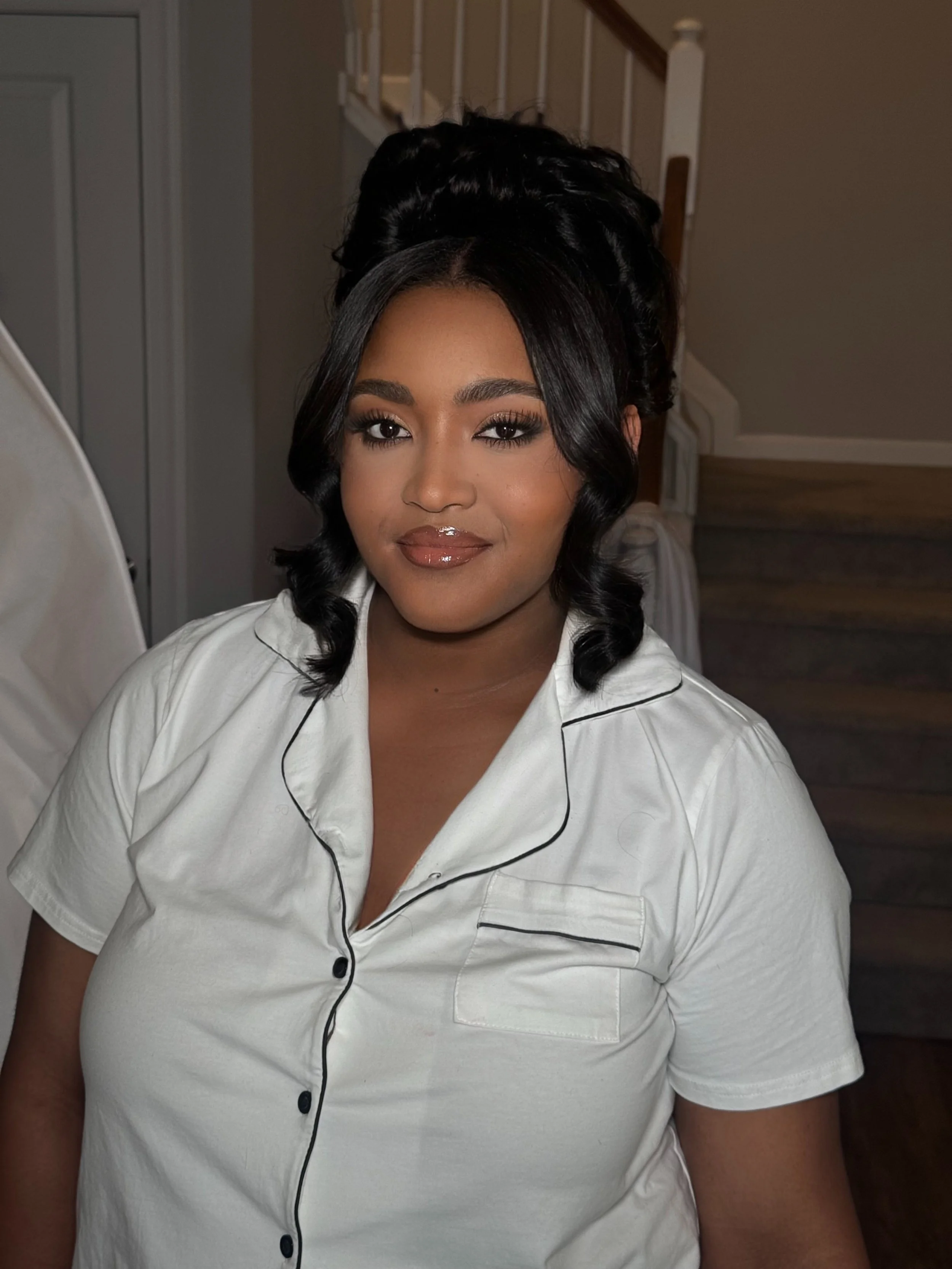 A woman with black, wavy hair wearing a white, short-sleeved pajama shirt with black piping, sitting indoors near a staircase.
