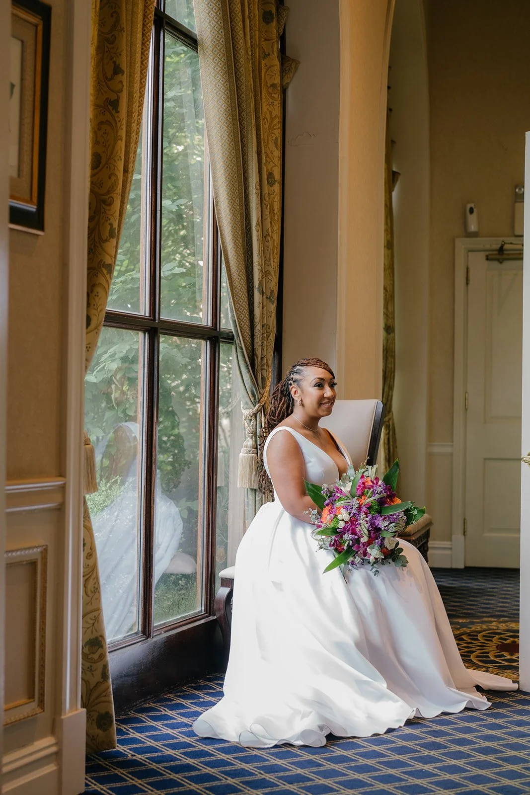 A bride sitting on a chair holding a colorful bouquet of flowers, next to a large window with gold curtains, in an elegant room with patterned carpet and cream walls.