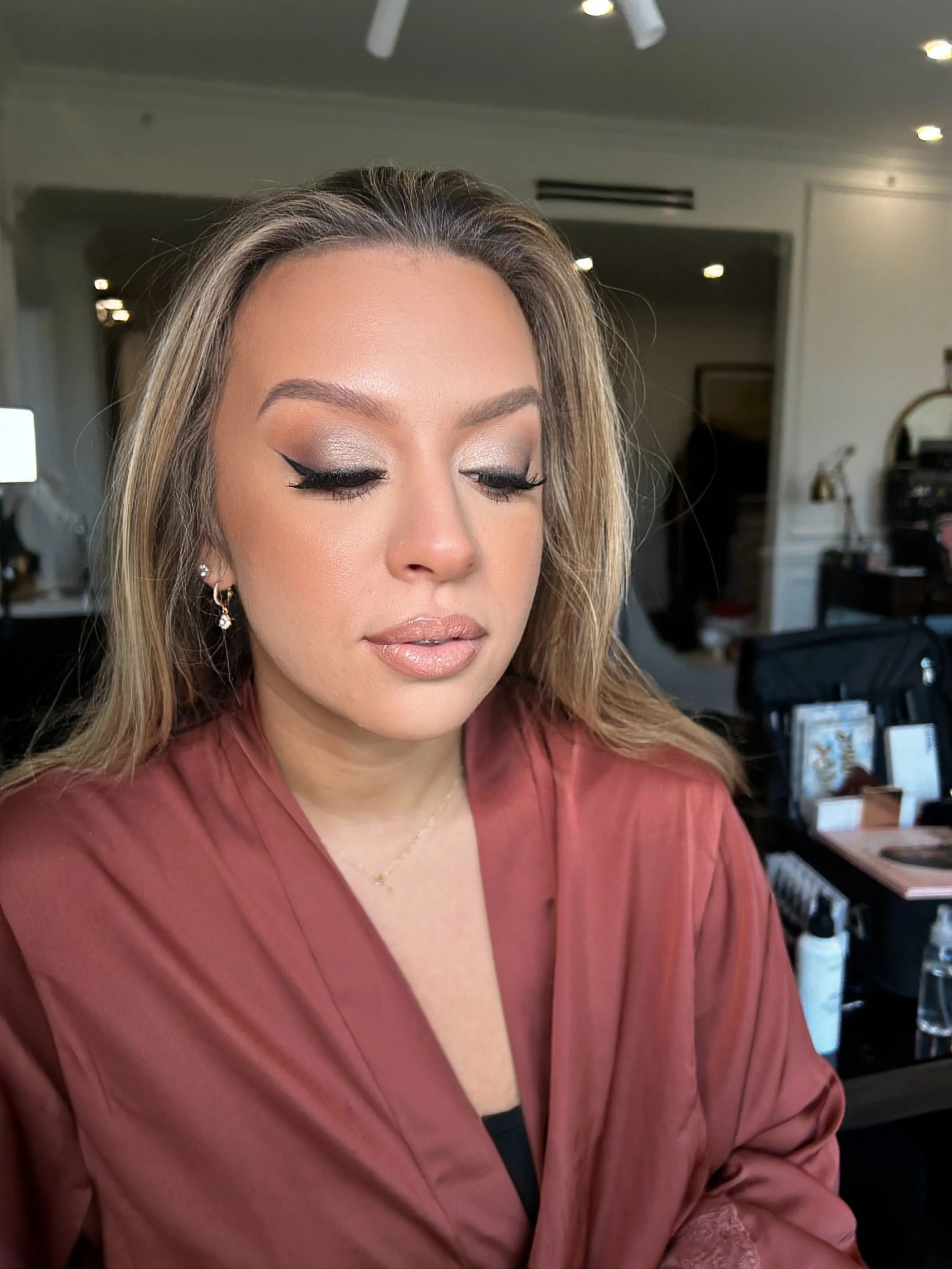 A woman with makeup, including winged eyeliner and nude lipstick, looks down with her eyes closed. She is wearing earrings and a rose-colored satin blouse, with a background of a room containing various objects and furniture.