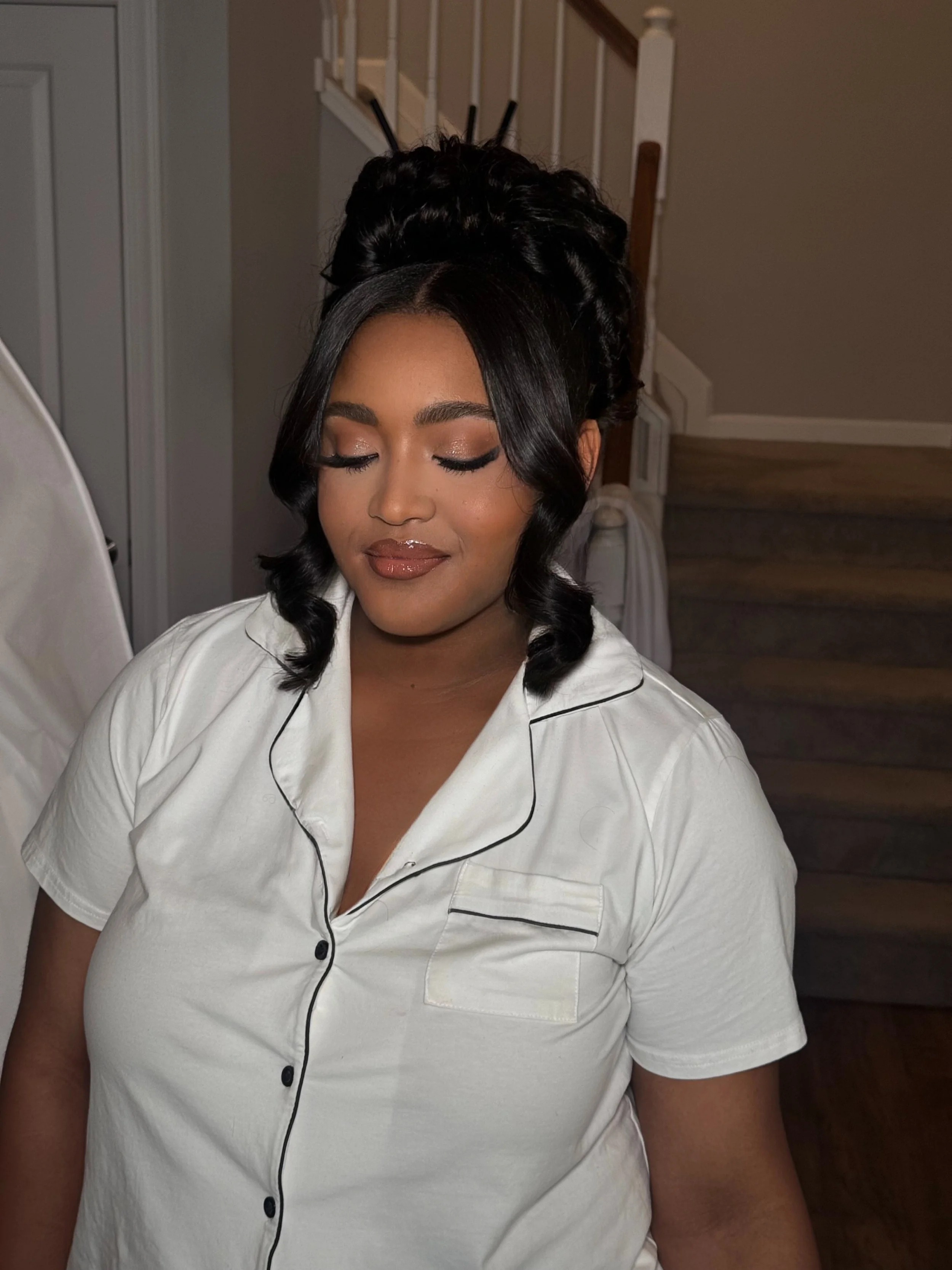 A woman with dark hair in curled waves and an intricate updo, wearing a white satin pajama top with black piping, standing in a dimly lit room with stairs in the background, her eyes closed and a calm expression.