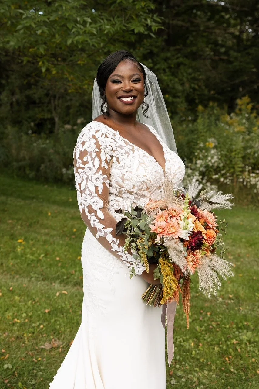 A smiling bride standing outdoors in a white lace wedding dress with floral embroidery, holding a bouquet of pink, peach, maroon, and green flowers with greenery and pampas grass, with trees and bushes in the background.