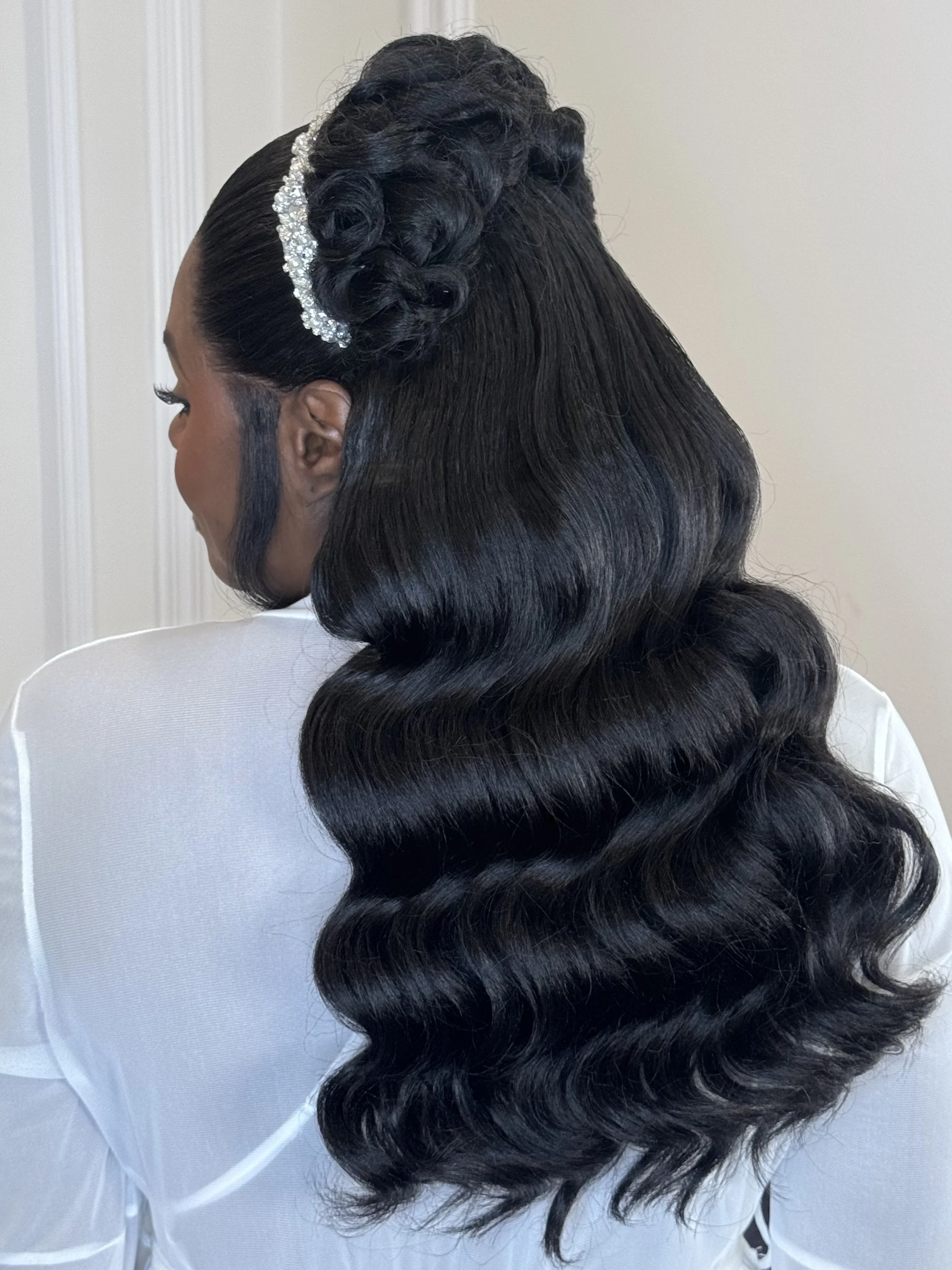 A woman's hairstyle featuring vintage-style glossy black waves with a side part, accessorized with a rhinestone headband.