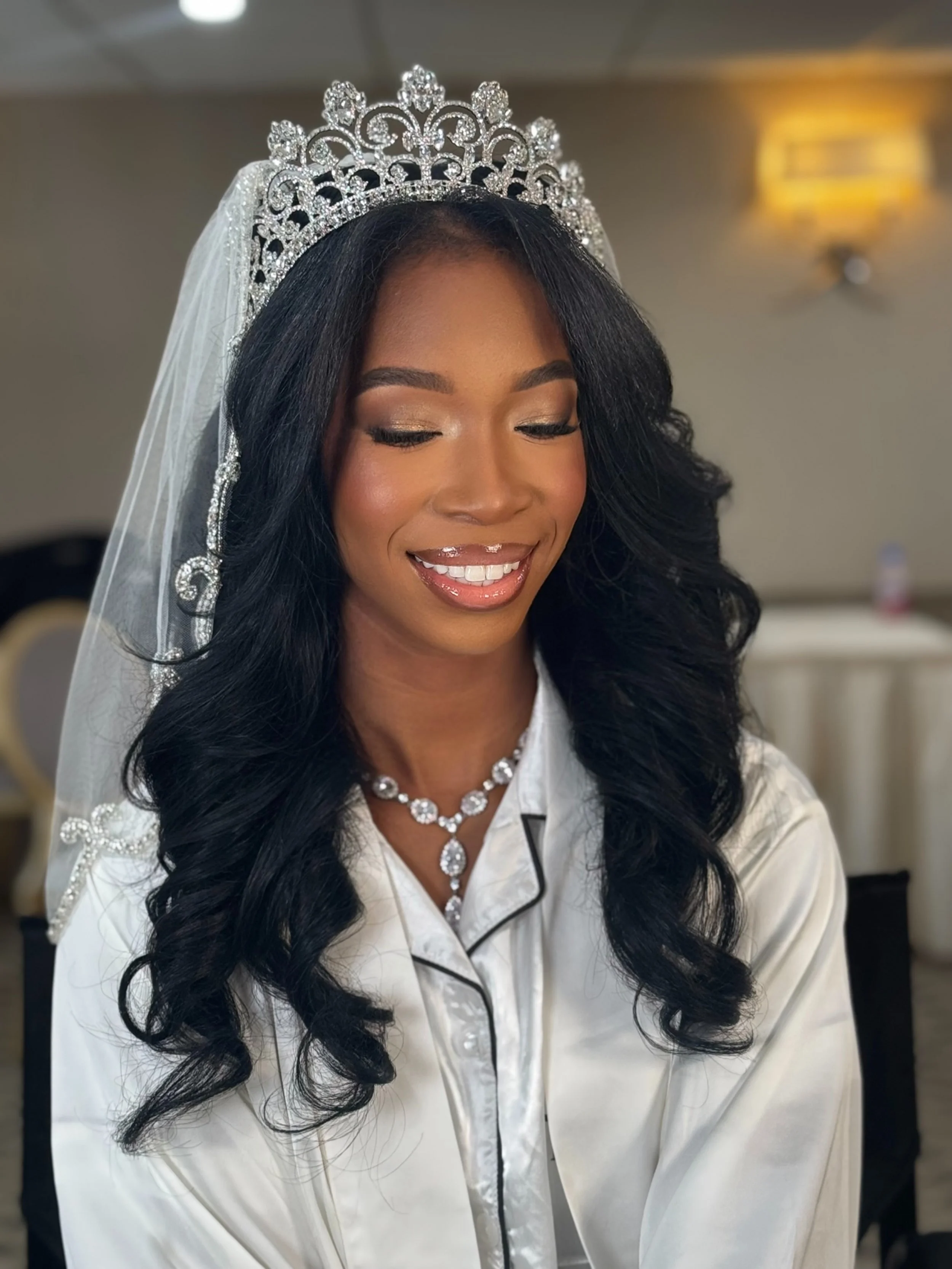 A woman with long, wavy black hair wearing a tiara, makeup, and a pearl necklace, smiling with her eyes closed in a dressing room.