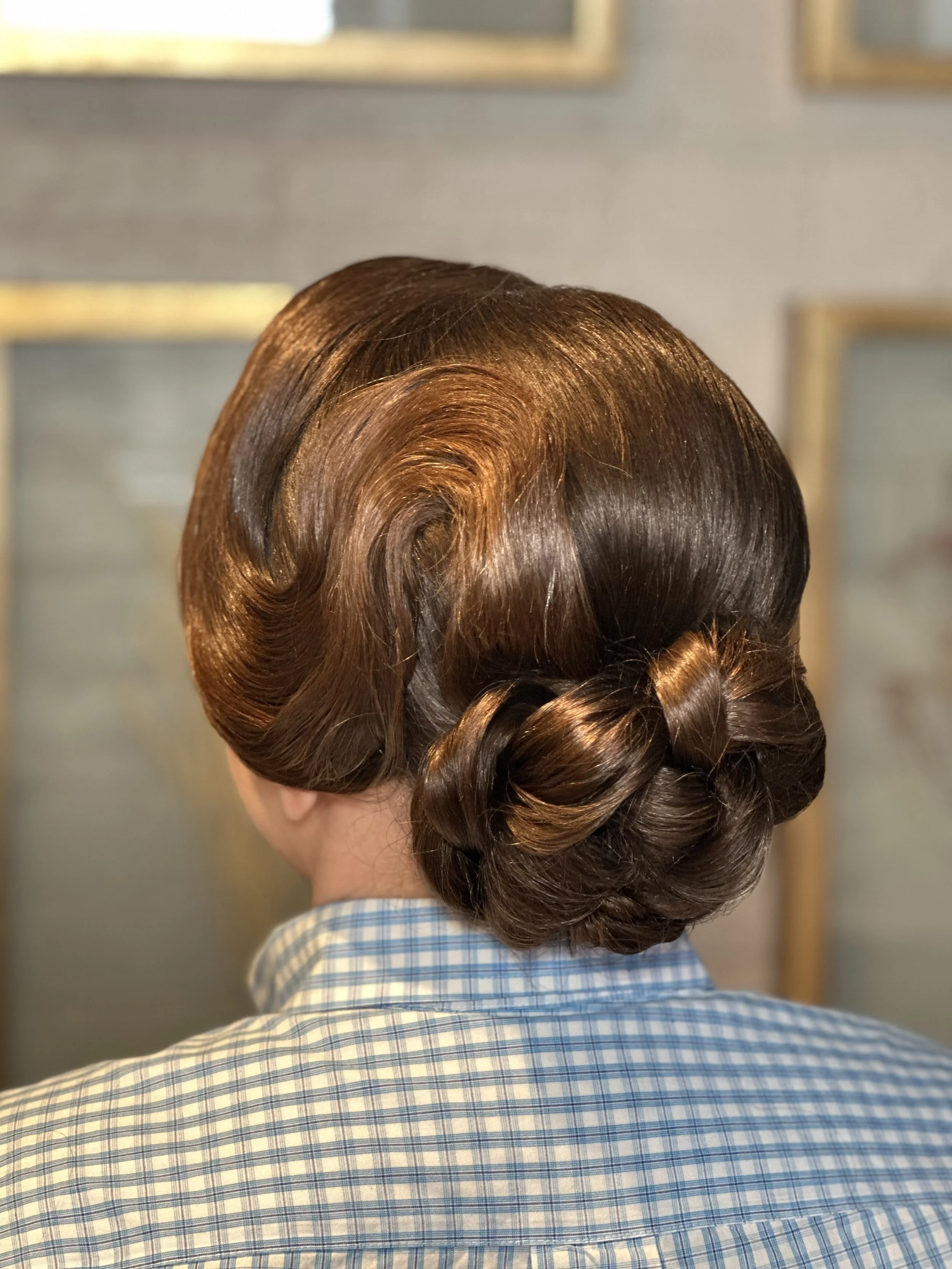 Back of a person's head with vintage styled brown hair, styled in large finger waves and a braid, wearing a blue and white checkered shirt.