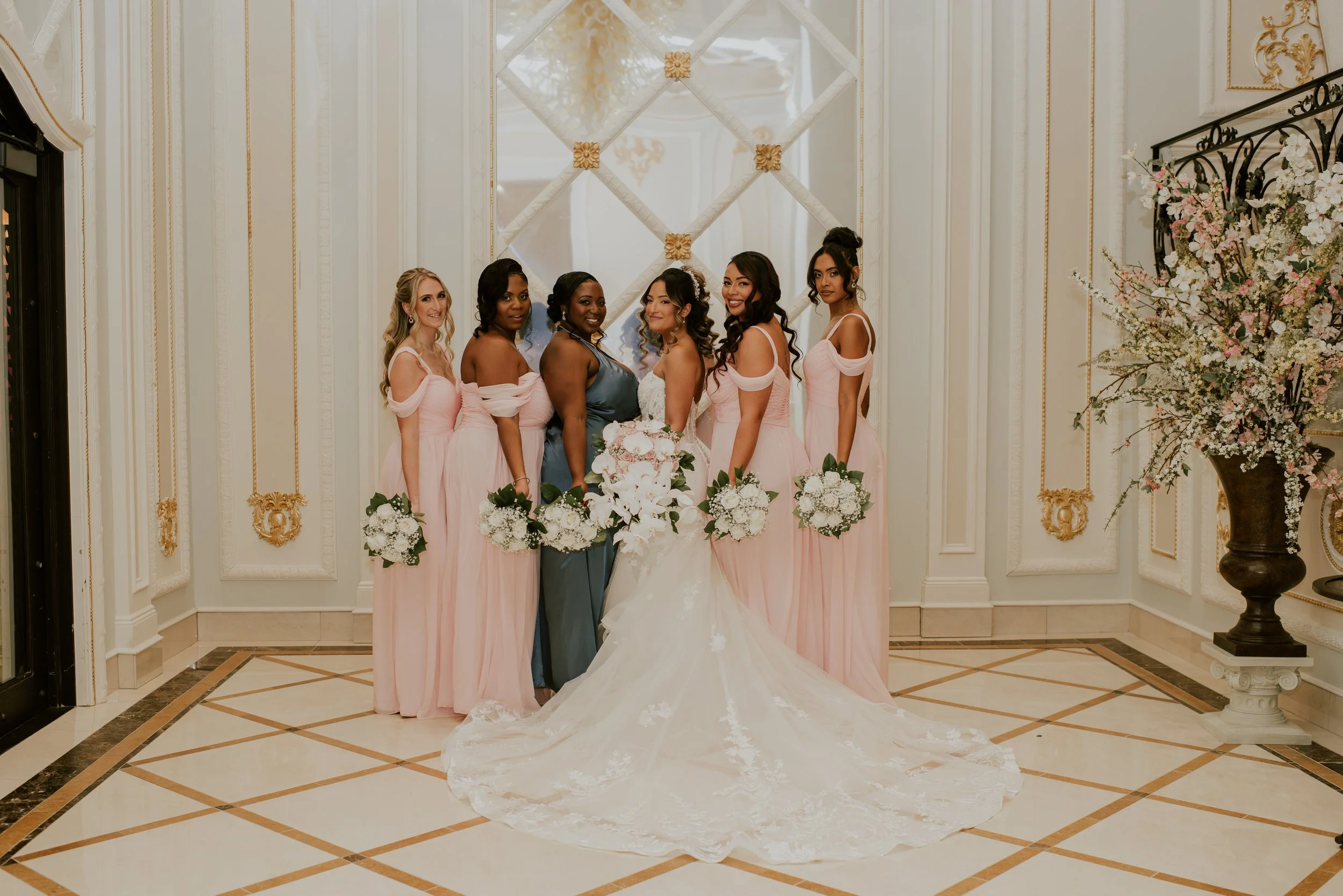 A group of six women in pink bridesmaid dresses and one woman in a white wedding gown, standing together in an elegant decorated room for a wedding celebration.