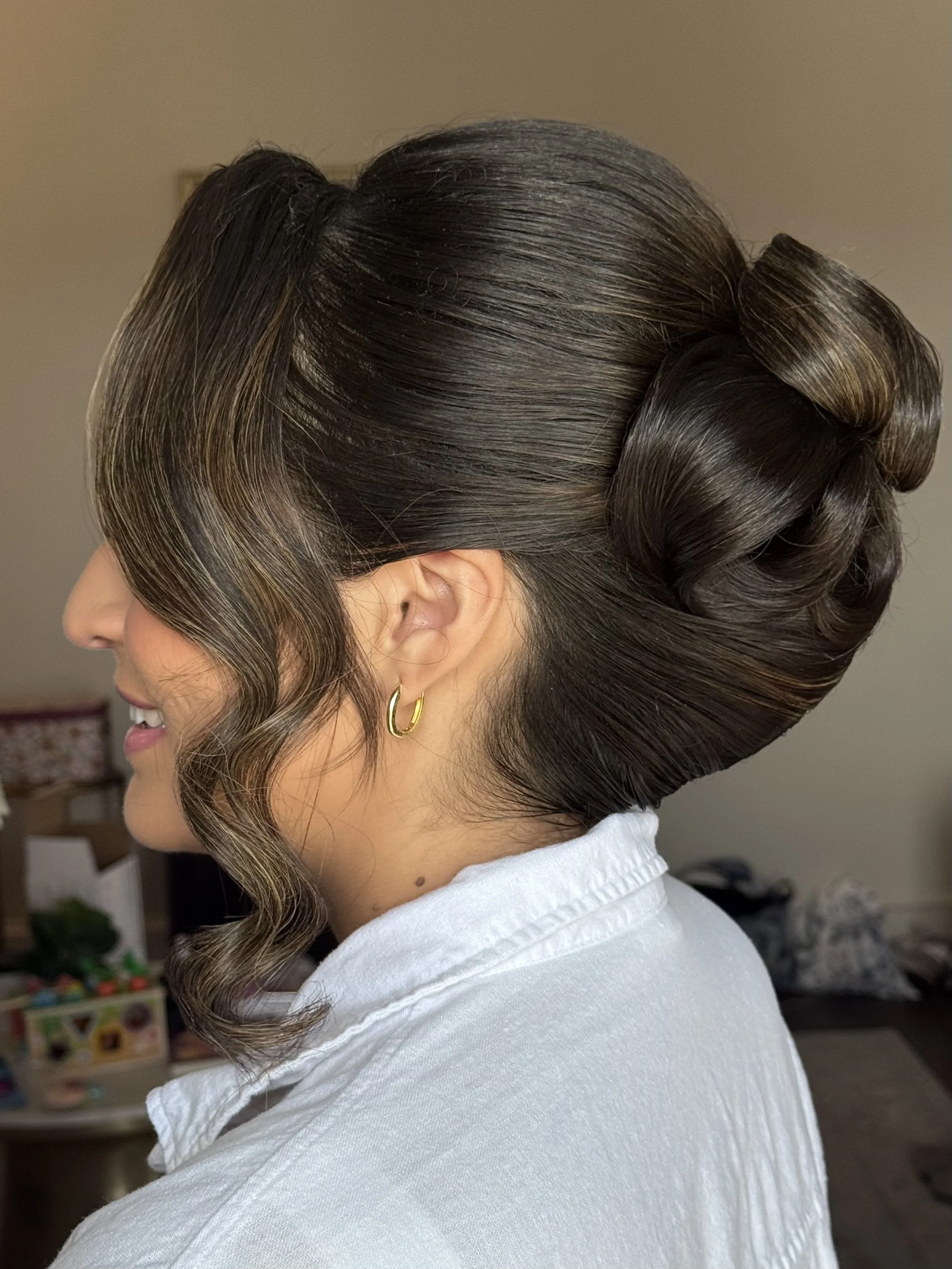 A woman with dark brown, voluminous, vintage-inspired hairstyle with curled ends and a deep side part, wearing a white top and gold hoop earrings.