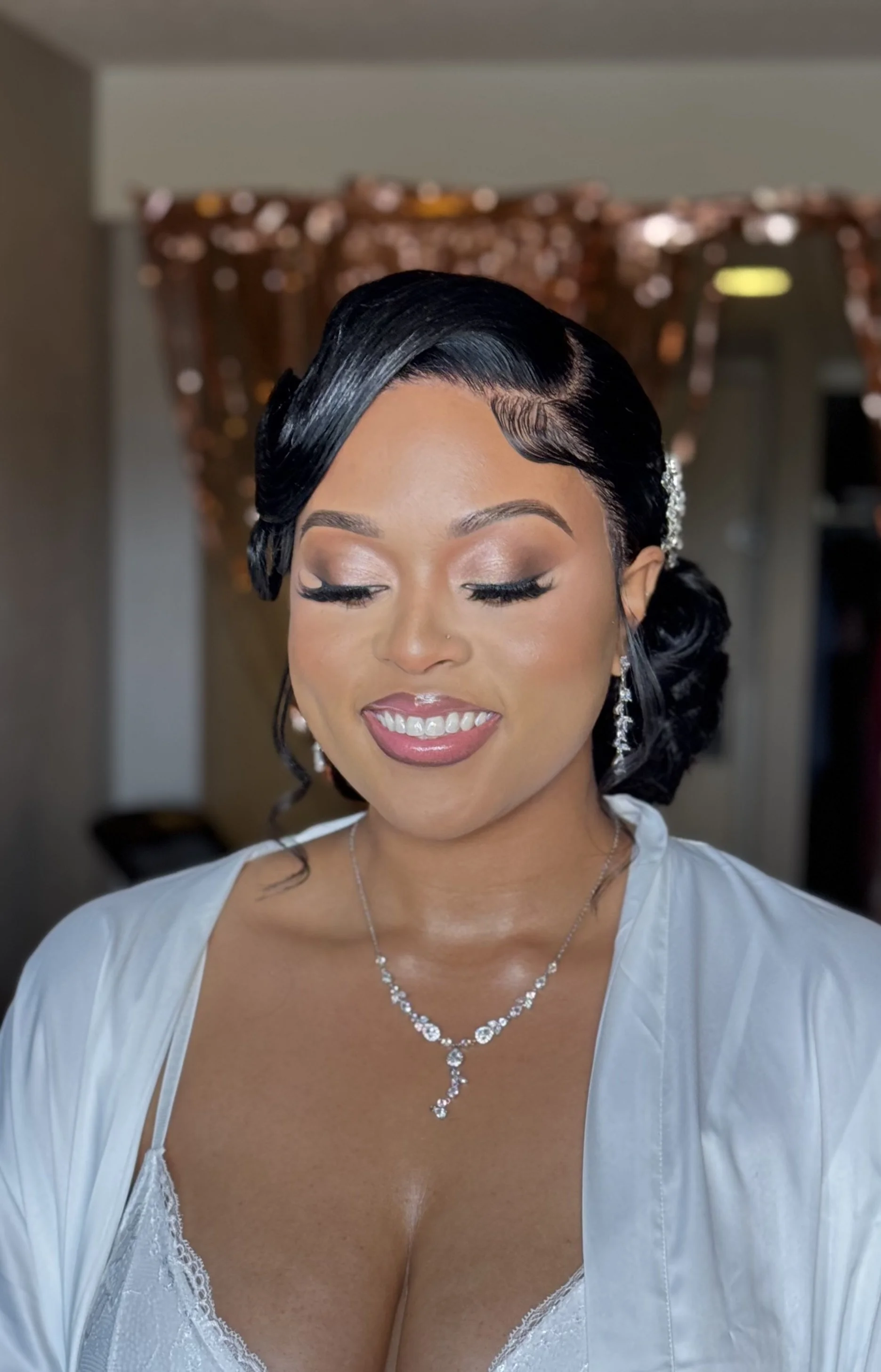 A woman with styled black hair, makeup, and jewelry, wearing a white satin robe and lace lingerie, smiling with eyes closed in an indoor setting.