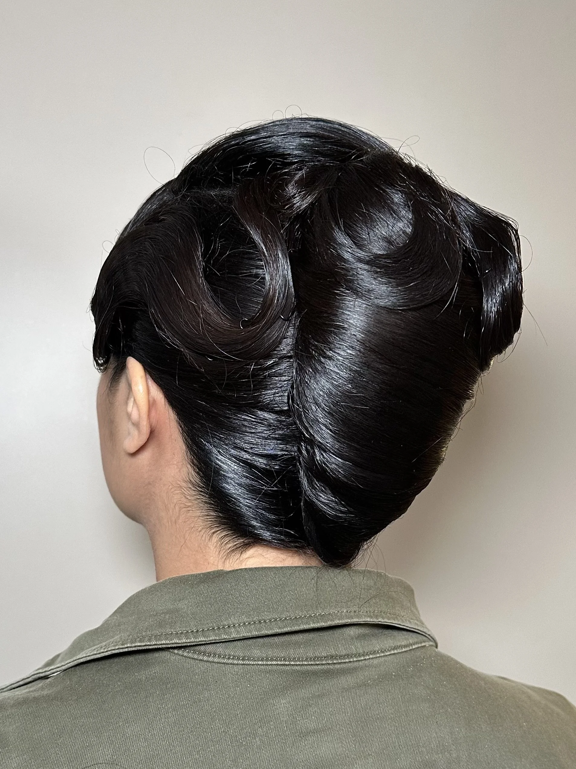 Back of a person's head with vintage, dark, shiny, slicked-back hair, wearing a green shirt.
