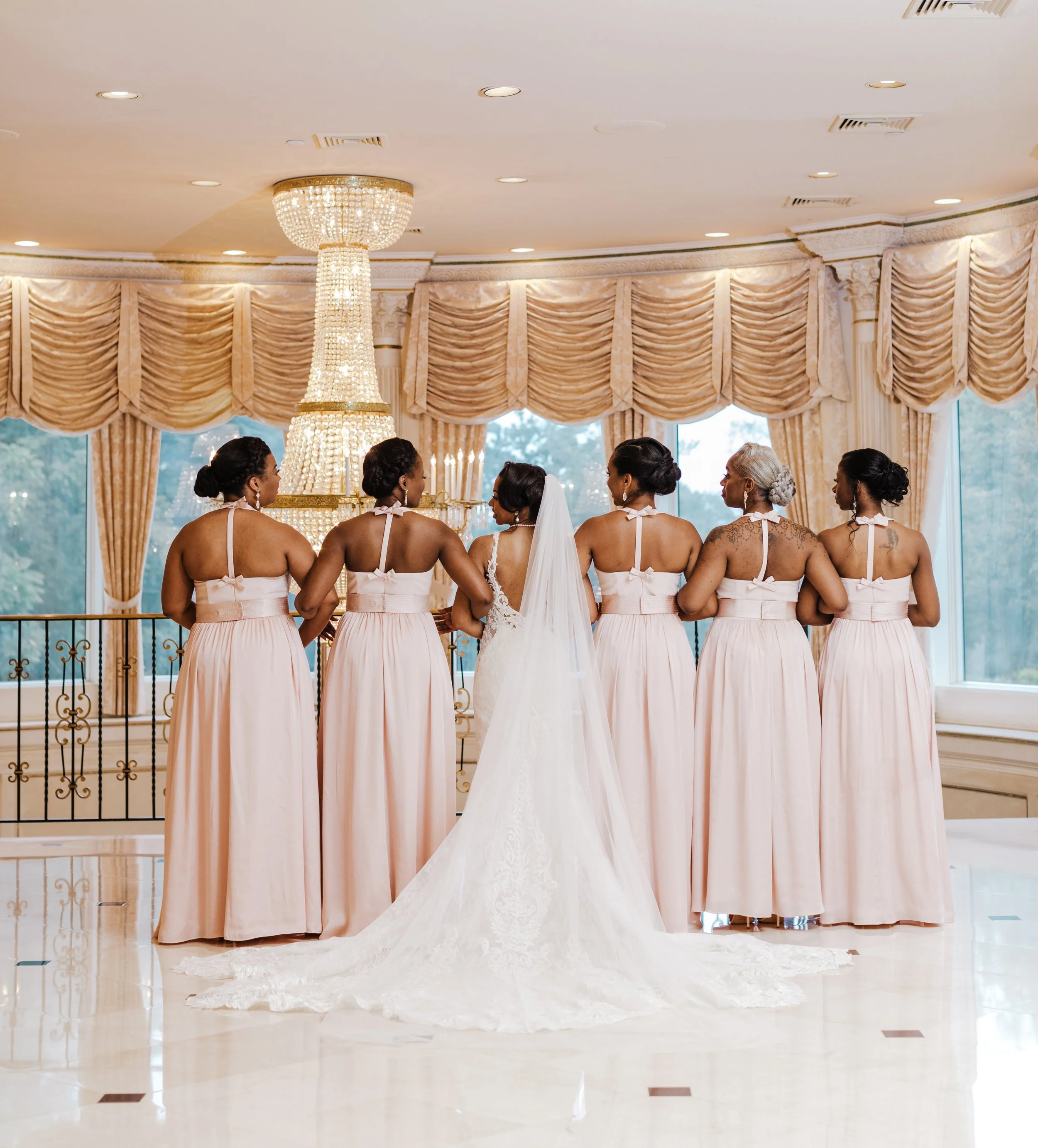 A group of women in matching blush pink dresses with bow accents, standing hand in hand in a luxurious, decorated room with a chandelier and gathered curtains, likely preparing for a wedding.