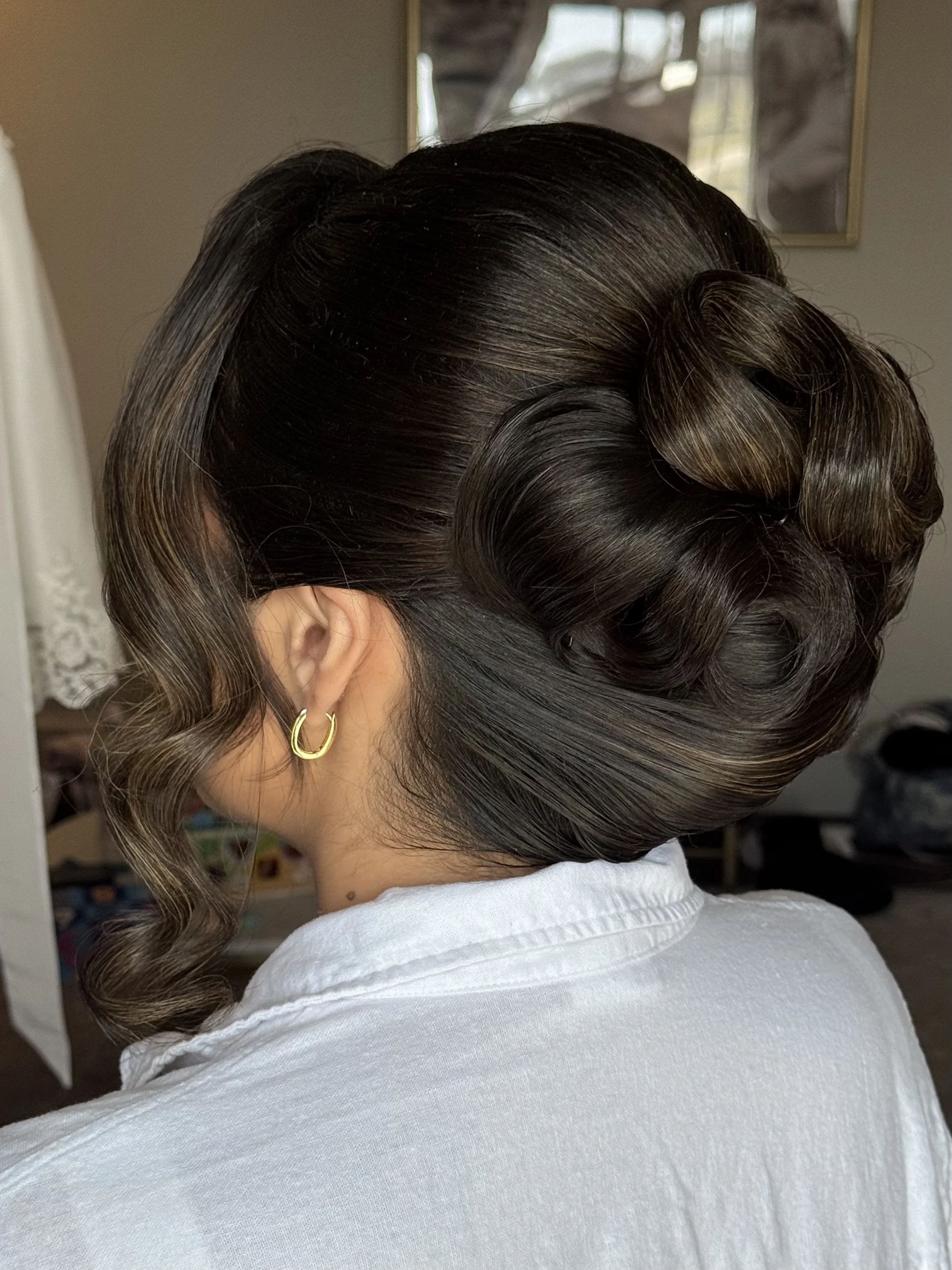 A woman with dark and light brown curled hair styled in an elegant updo, wearing gold hoop earrings, and dressed in a white shirt.