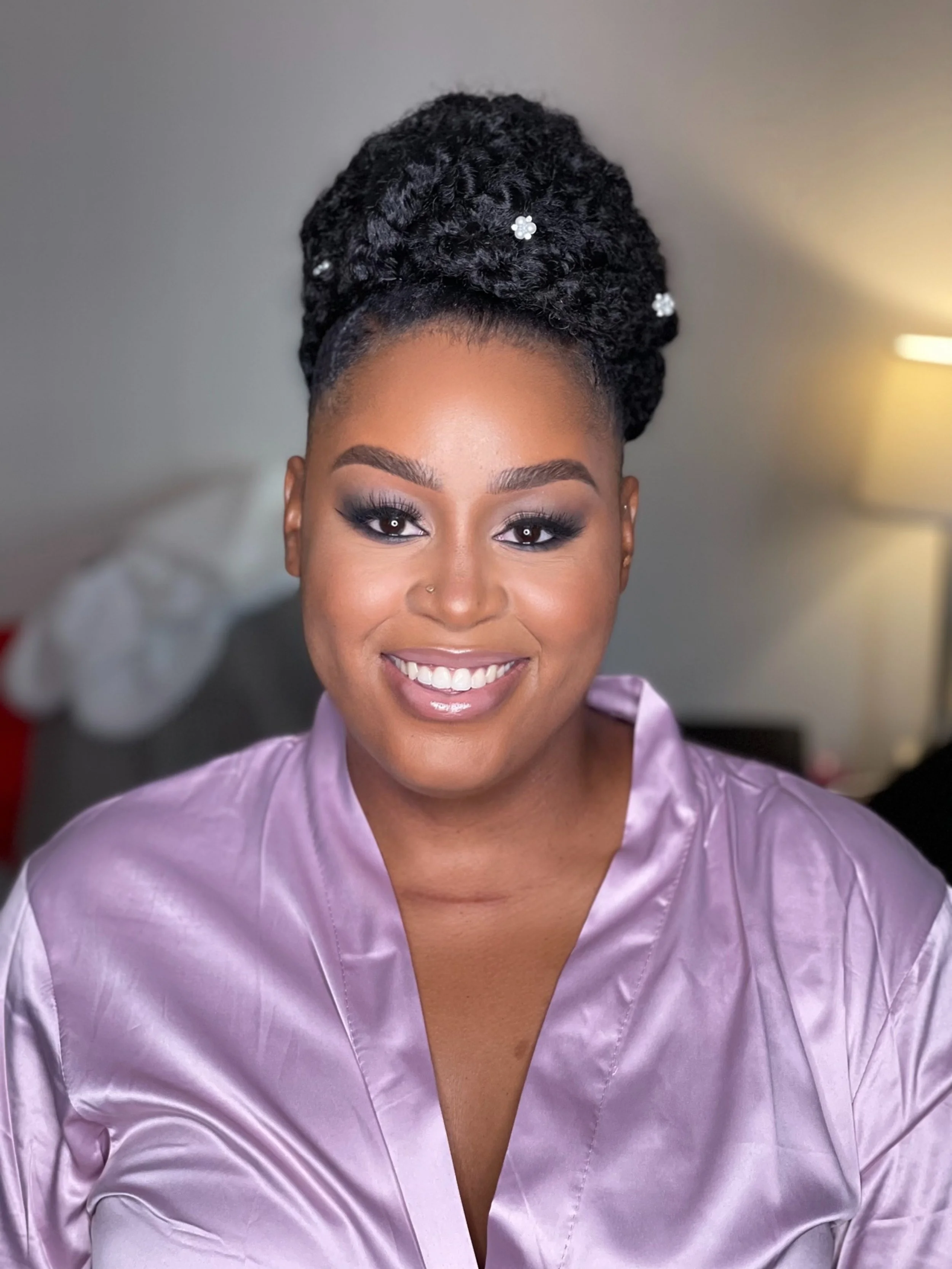 Close-up photo of a woman with styled black hair adorned with small white floral hairpins, wearing makeup with dark eyeshadow, and a light purple satin robe, smiling.