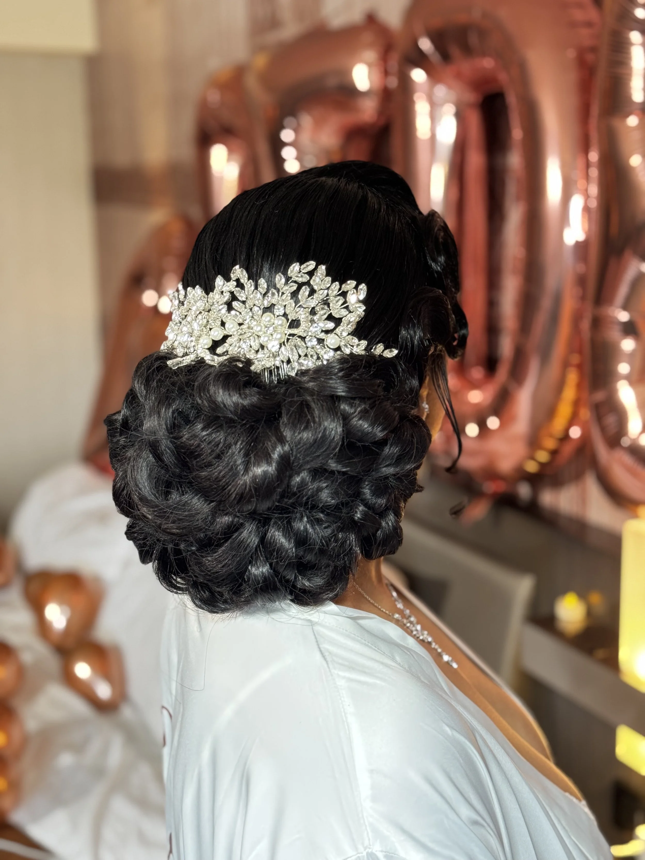 Close-up of a woman with black, curled hair styled in an elegant updo adorned with a large, intricate silver and pearl hair accessory. She is wearing a white dress with a pearl necklace. The background features decorative, metallic balloon numbers an