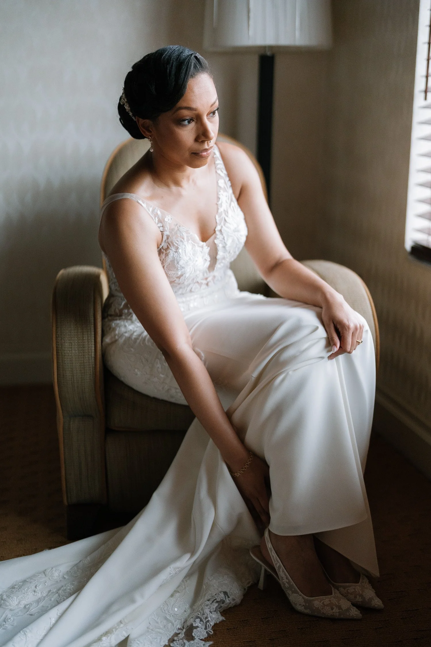 A woman in a wedding dress sitting on a chair, adjusting her shoe, with her dress extended on the floor. She has dark hair styled in an updo and is near a window with blinds, in an indoor setting.