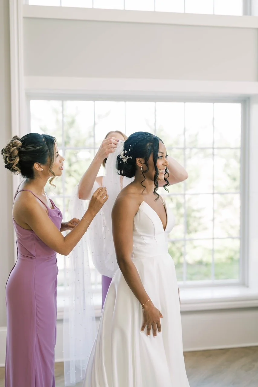 A bride in a white wedding dress is smiling as her bridesmaids assist her with her veil in a bright room with large windows.