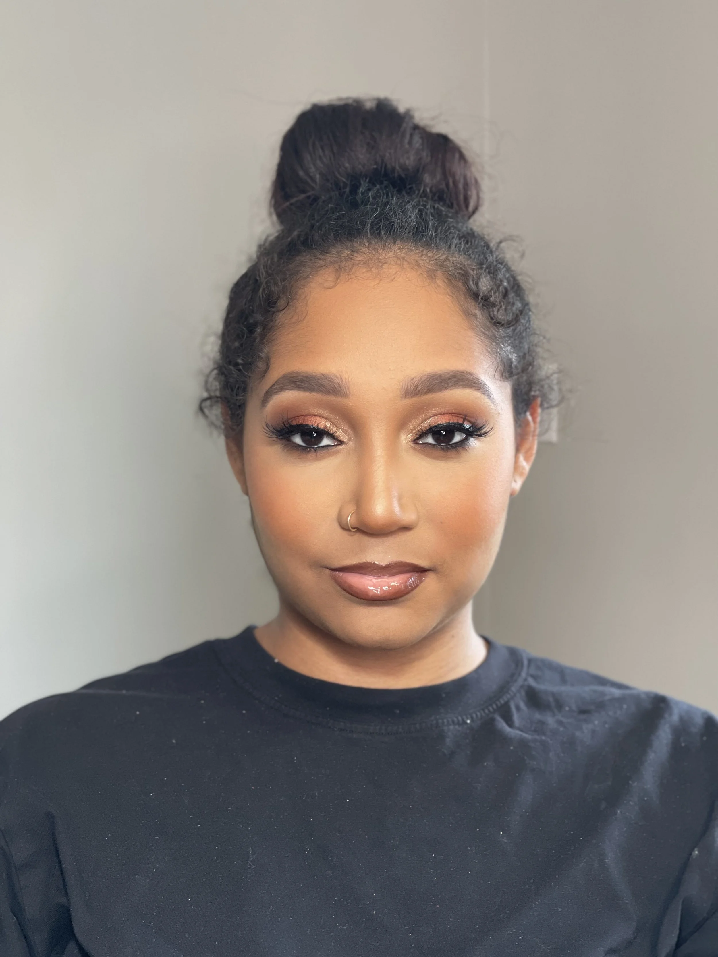 Close-up of a woman with styled hair in a high bun, makeup, and a nose ring, wearing a black shirt.