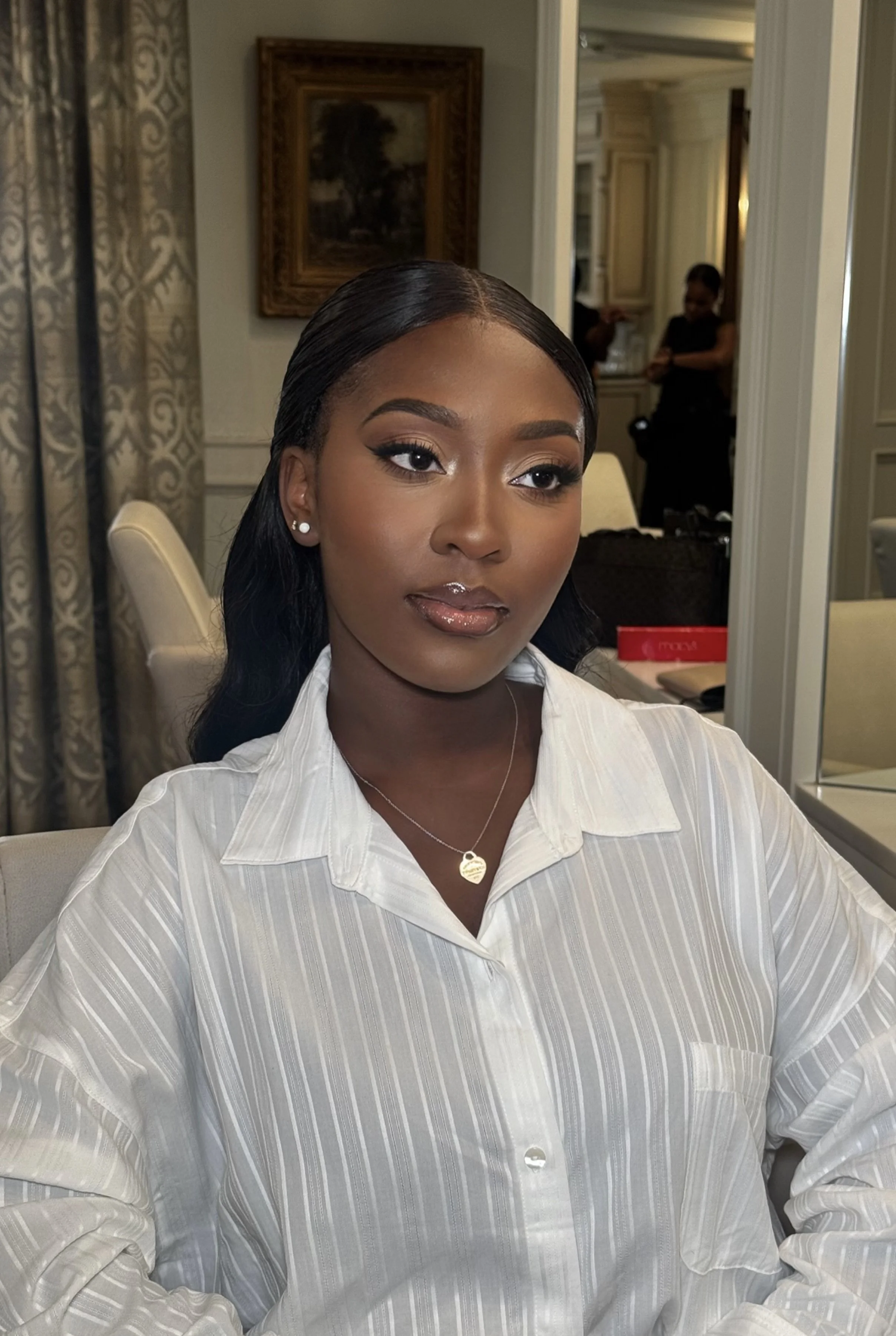 A woman with dark hair and makeup, wearing pearl earrings, a gold heart necklace, and a white striped shirt, sitting indoors with a mirror and two women in black outfits in the background.