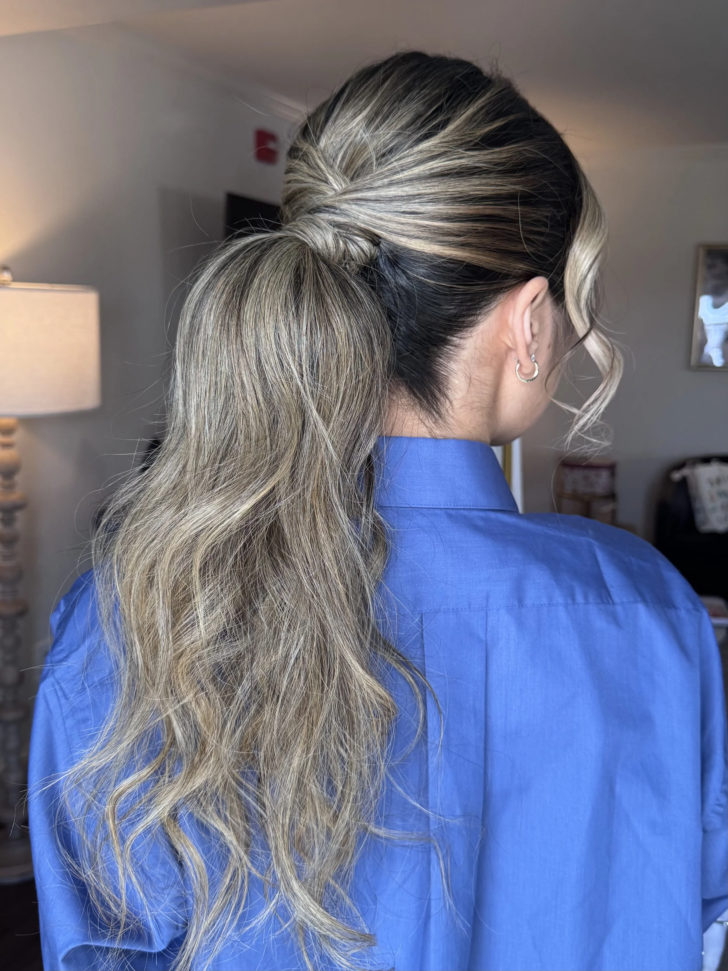 A woman with long, wavy blonde hair styled in a loose ponytail, wearing a blue shirt, in a room with a lamp and framed picture on the wall.