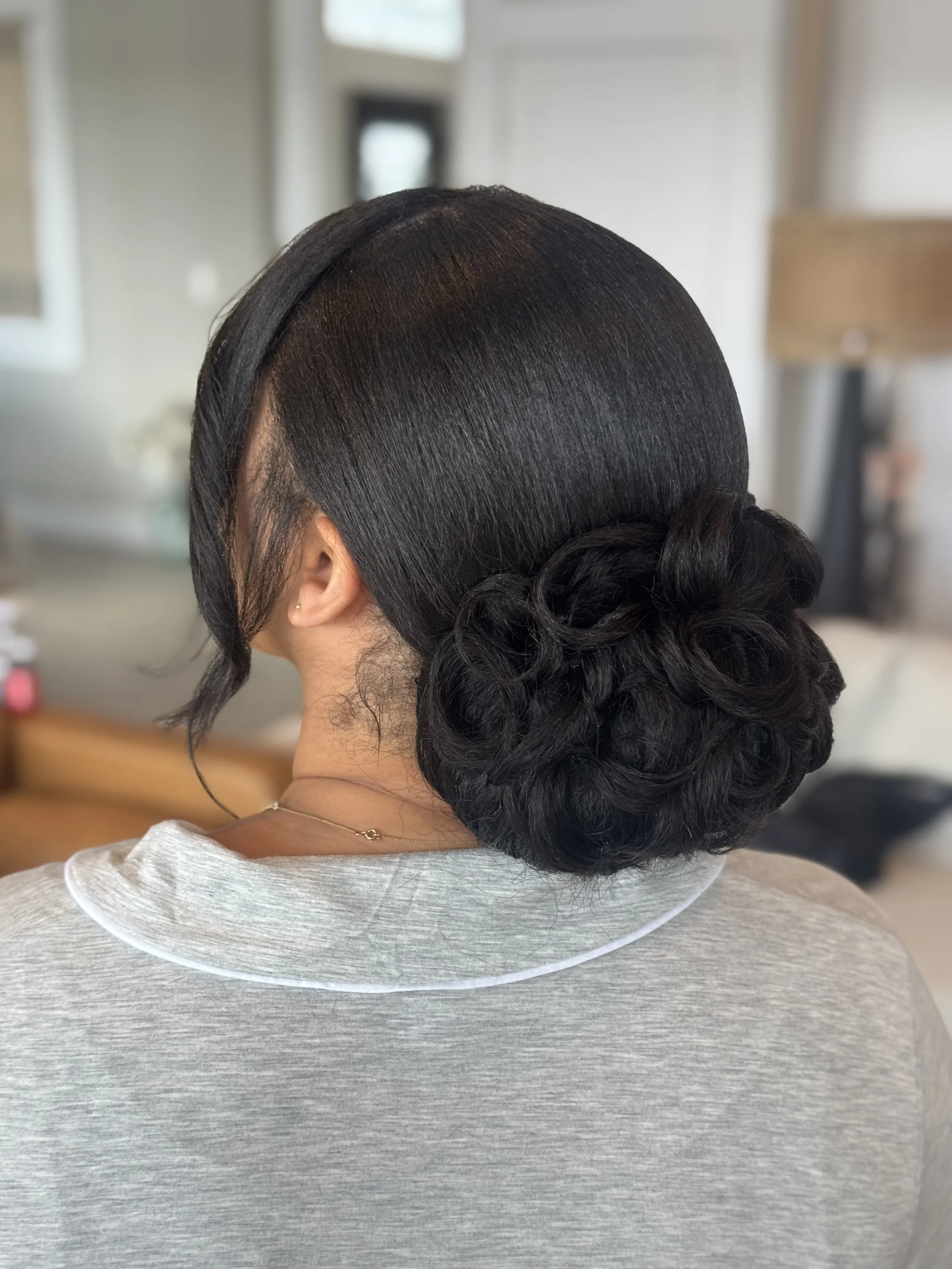 A woman with dark, shiny, straight hair styled in a low bun with a side part, wearing a light gray shirt and a delicate necklace, sitting at a table in a room with neutral decor.
