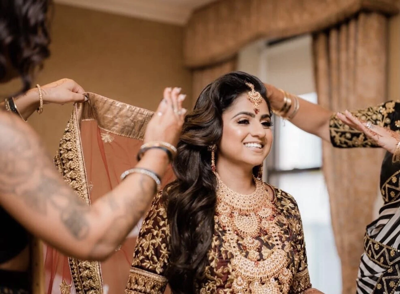 A woman dressed in traditional Indian bridal attire being assisted by women in ornate clothing, as they help her with her outfit or jewelry.
