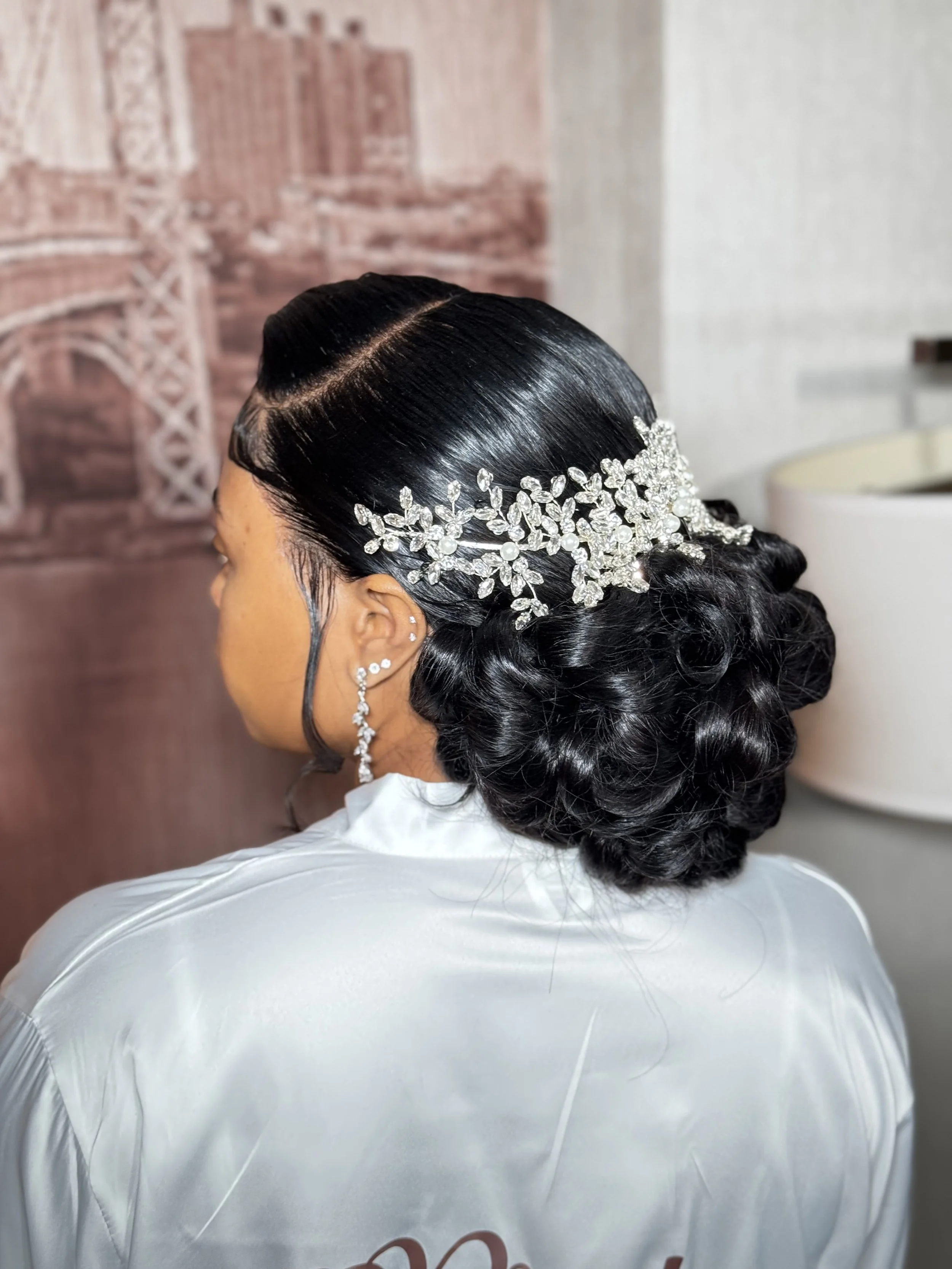 Side view of a woman with glossy black hair styled in loose curls, wearing a silver rhinestone and pearl hairpiece, earrings, and a white satin garment.