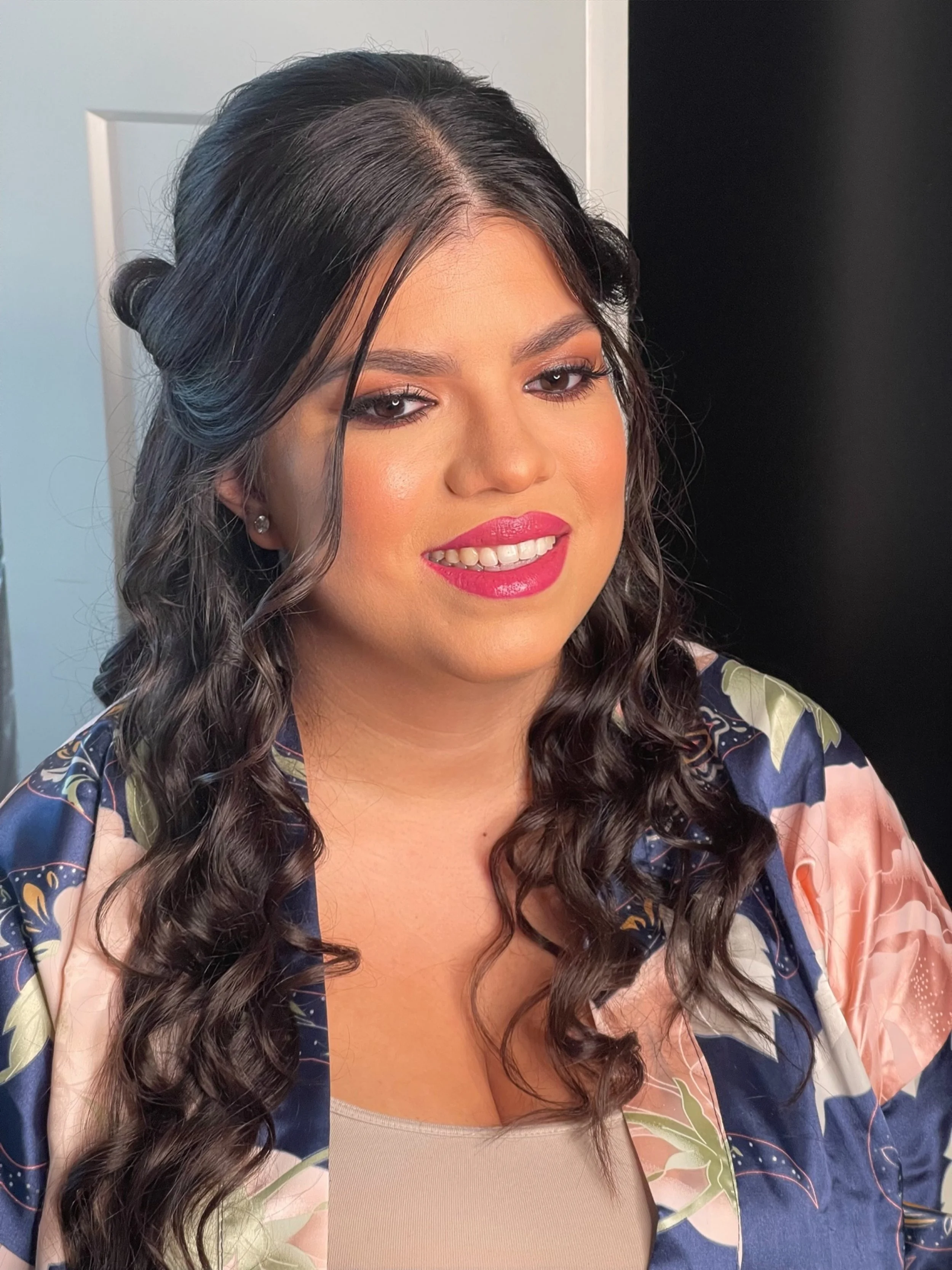 Close-up of a woman with curly dark hair, smiling with pink lipstick, wearing earrings, and a patterned silk robe over a beige top, against a neutral background.
