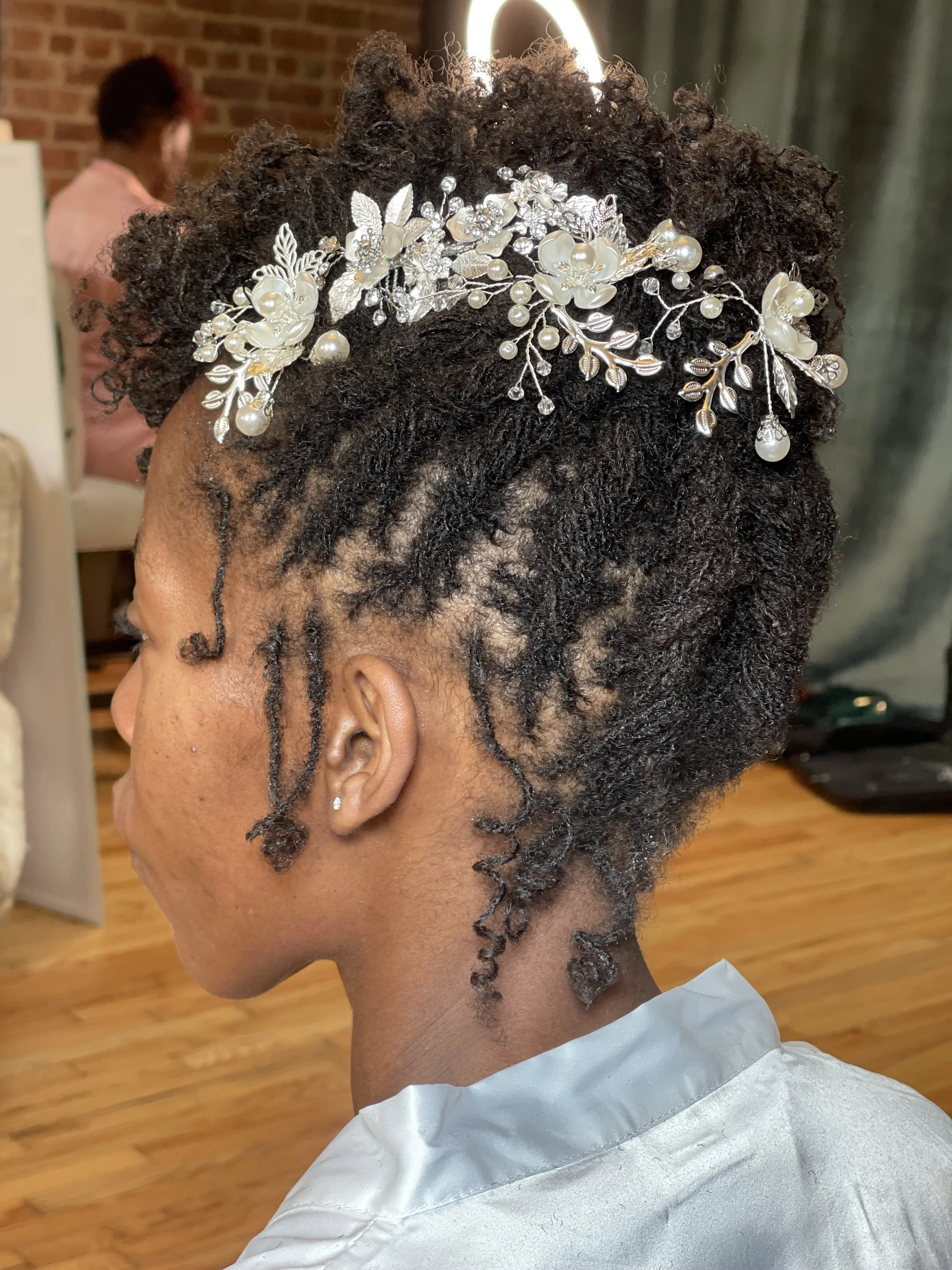Close-up of a woman with natural curly hair adorned with a silver floral hair accessory, in a salon with a woman in the background and a hardwood floor.